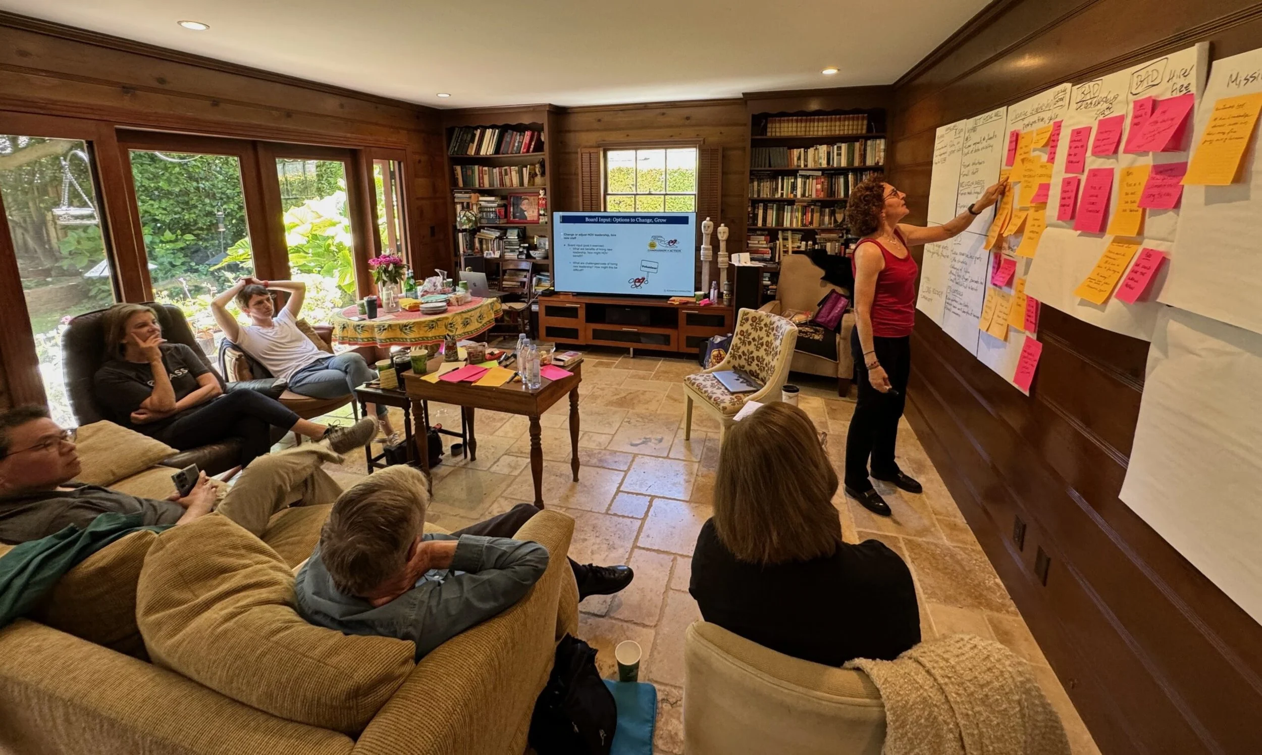 A woman presents in a room filled with seated people, a large wall with white paper and pink sticky notes, bookshelves, and a television screen.