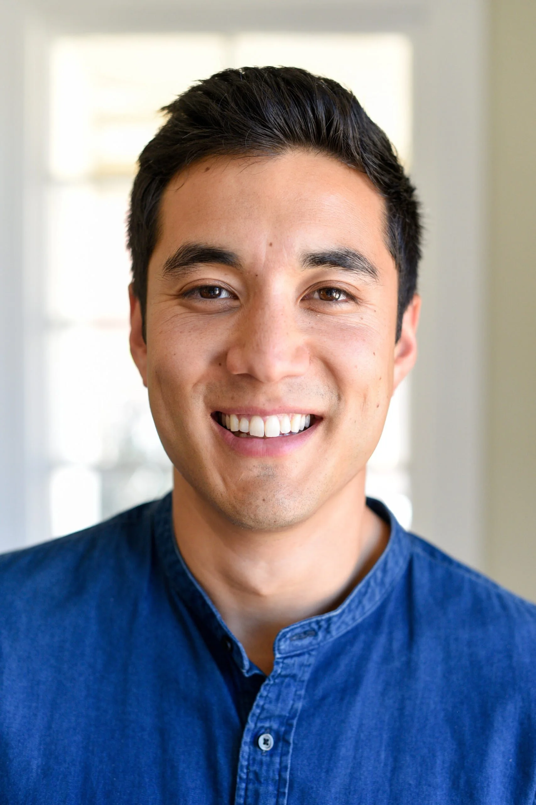 Close-up of a young man with short, dark hair smiling in a brightly lit room.