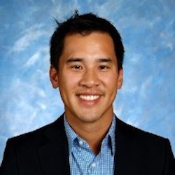 A smiling man with short black hair, wearing a black blazer over a blue and white patterned shirt, against a blue textured background.