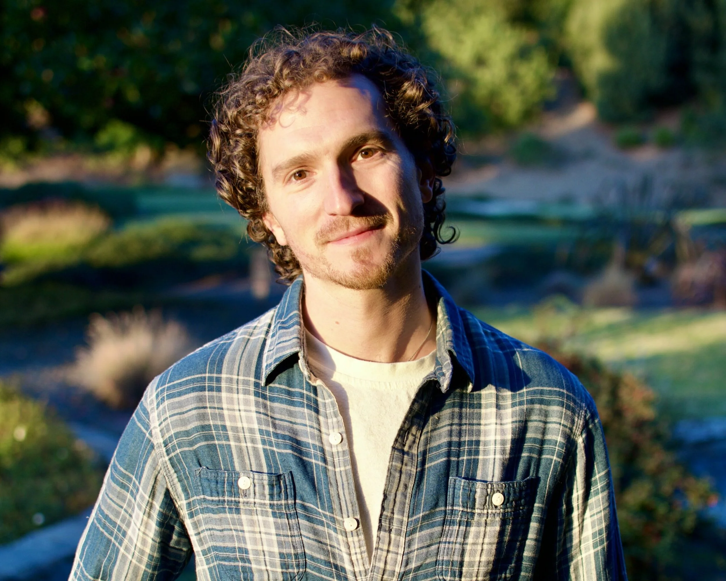 A young man with curly hair and a beard, wearing a plaid shirt and a light-colored T-shirt, standing outdoors with greenery and a river in the background during daytime.