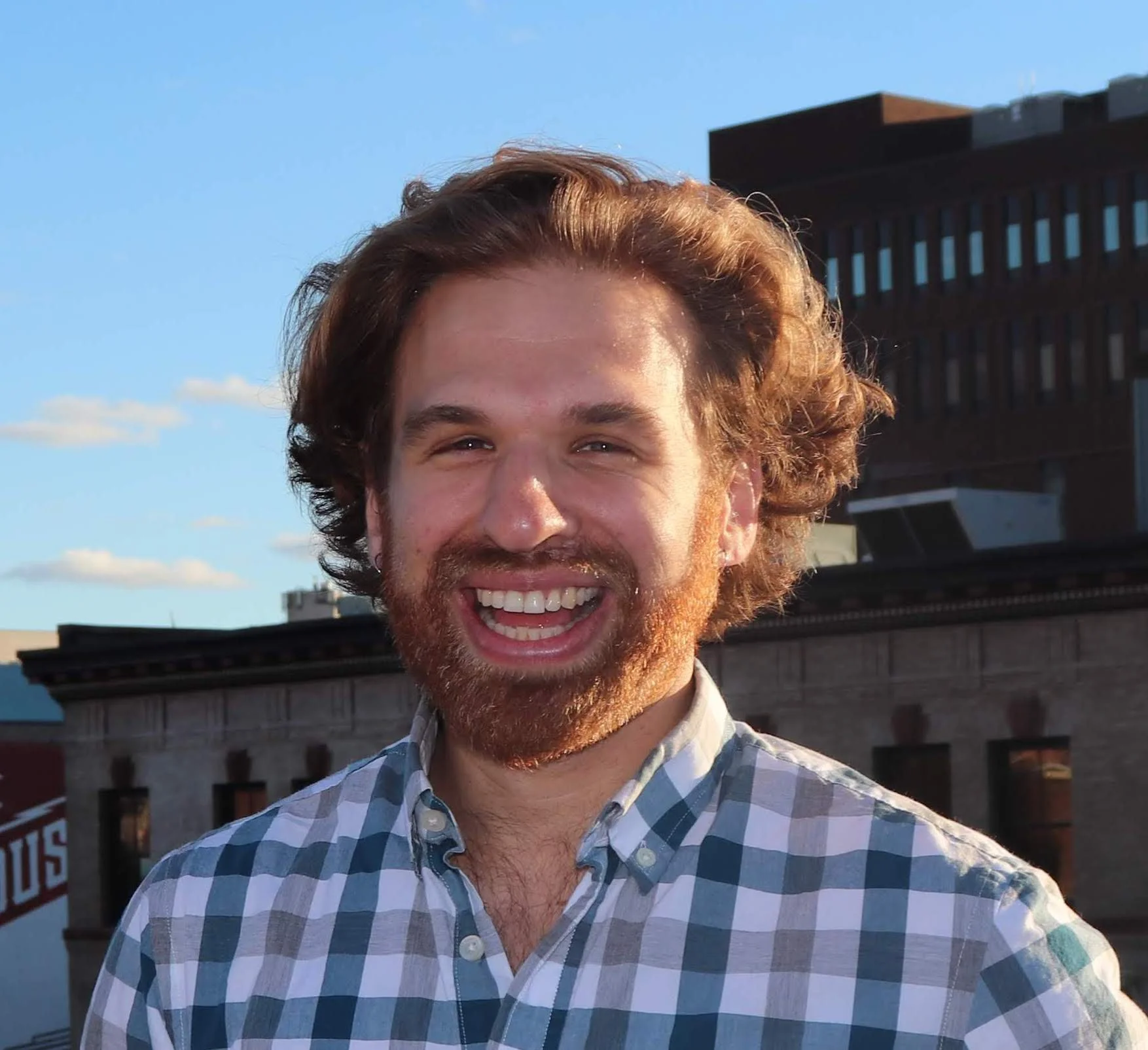 A smiling man with curly auburn hair and a beard, wearing a blue and white checkered shirt, standing outdoors in a city setting during the late afternoon or early evening.