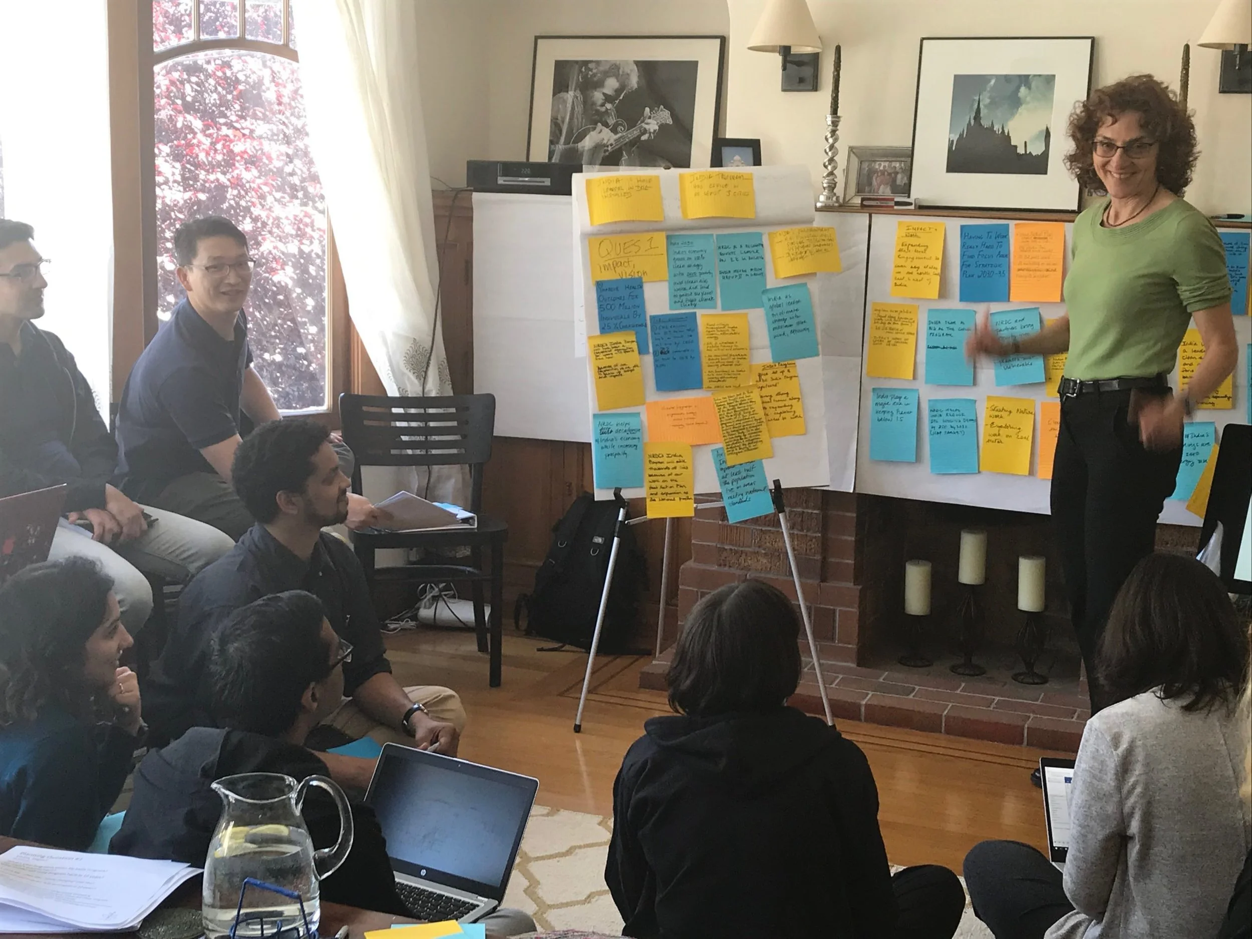A woman presenting in a room with six people seated on the floor, some with laptops. The woman is standing near large sheets of paper with blue and yellow sticky notes. The room has a fireplace with framed pictures and a window with curtains, letting in natural light.