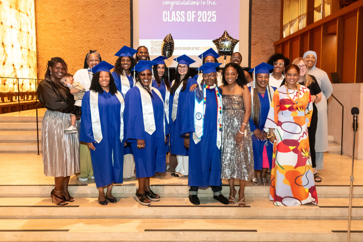 Group of graduates in blue caps and gowns posing for a photo on stage during a graduation ceremony, with some holding balloons and a large screen in the background that reads "congratulations to the class of 2025."