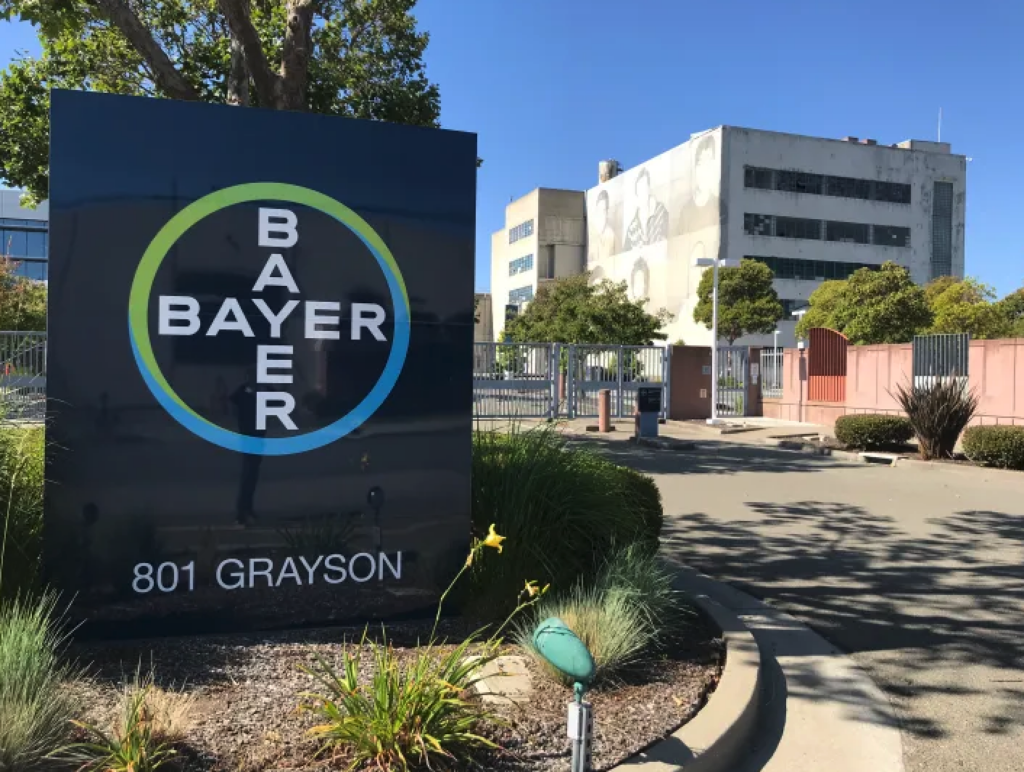 A Bayer office building at 801 Grayson, with a large sign in the foreground displaying the Bayer logo and address. The background shows a modern office building, trees, and a clear blue sky.