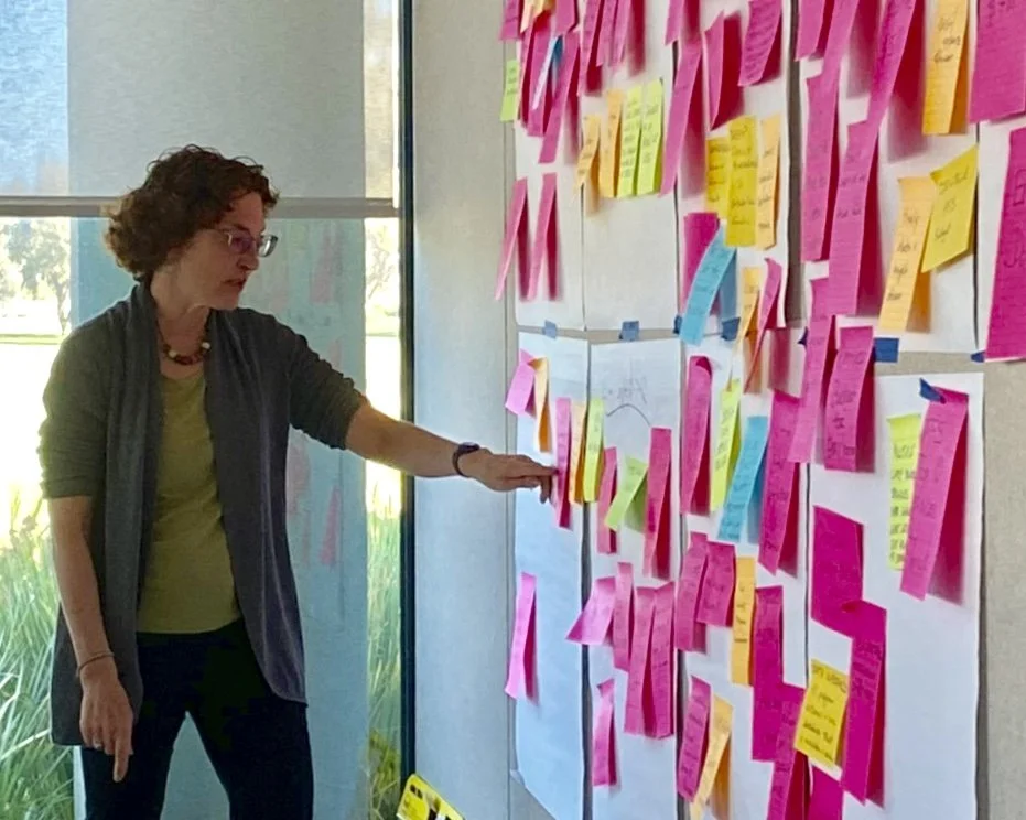 Woman placing a pink sticky note on a large wall covered with numerous colorful sticky notes during a brainstorming session.