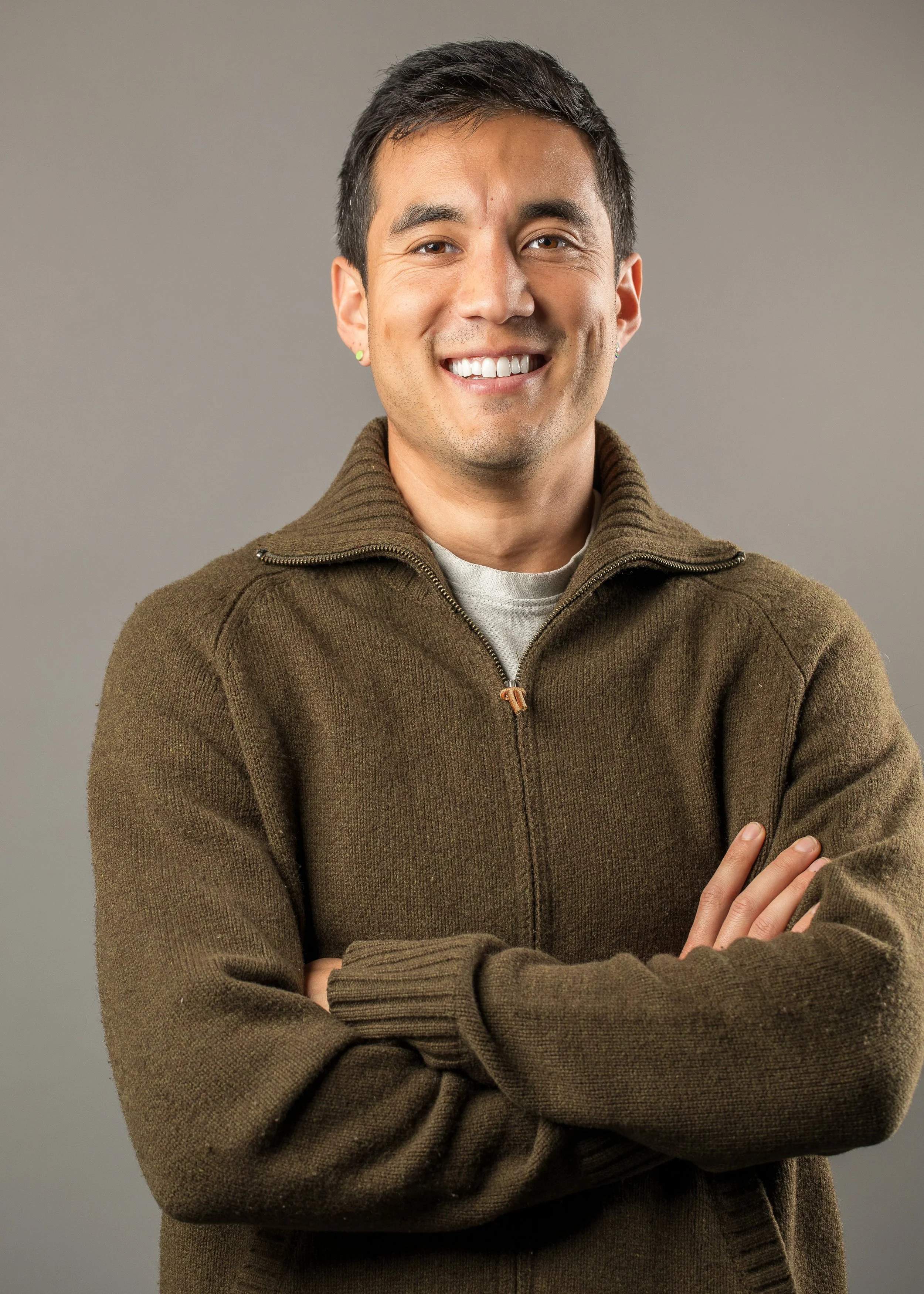 A smiling man with crossed arms, wearing a brown zip-up sweater and a light-colored shirt underneath, standing against a neutral gray background.