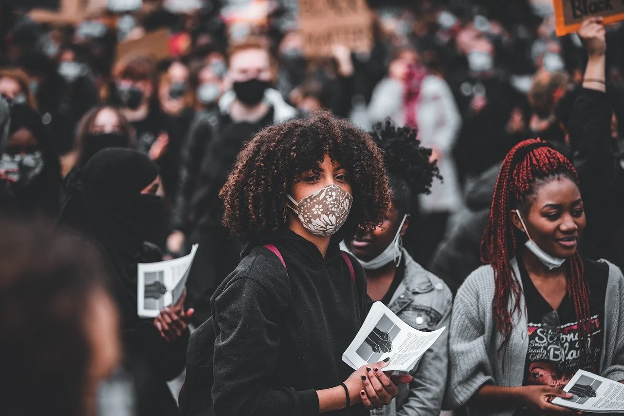 A group of people participating in a protest or rally, many wearing masks. The woman in the foreground has curly hair, is wearing a patterned face mask, and holds a pamphlet. Others around her also hold pamphlets, and some are holding signs.