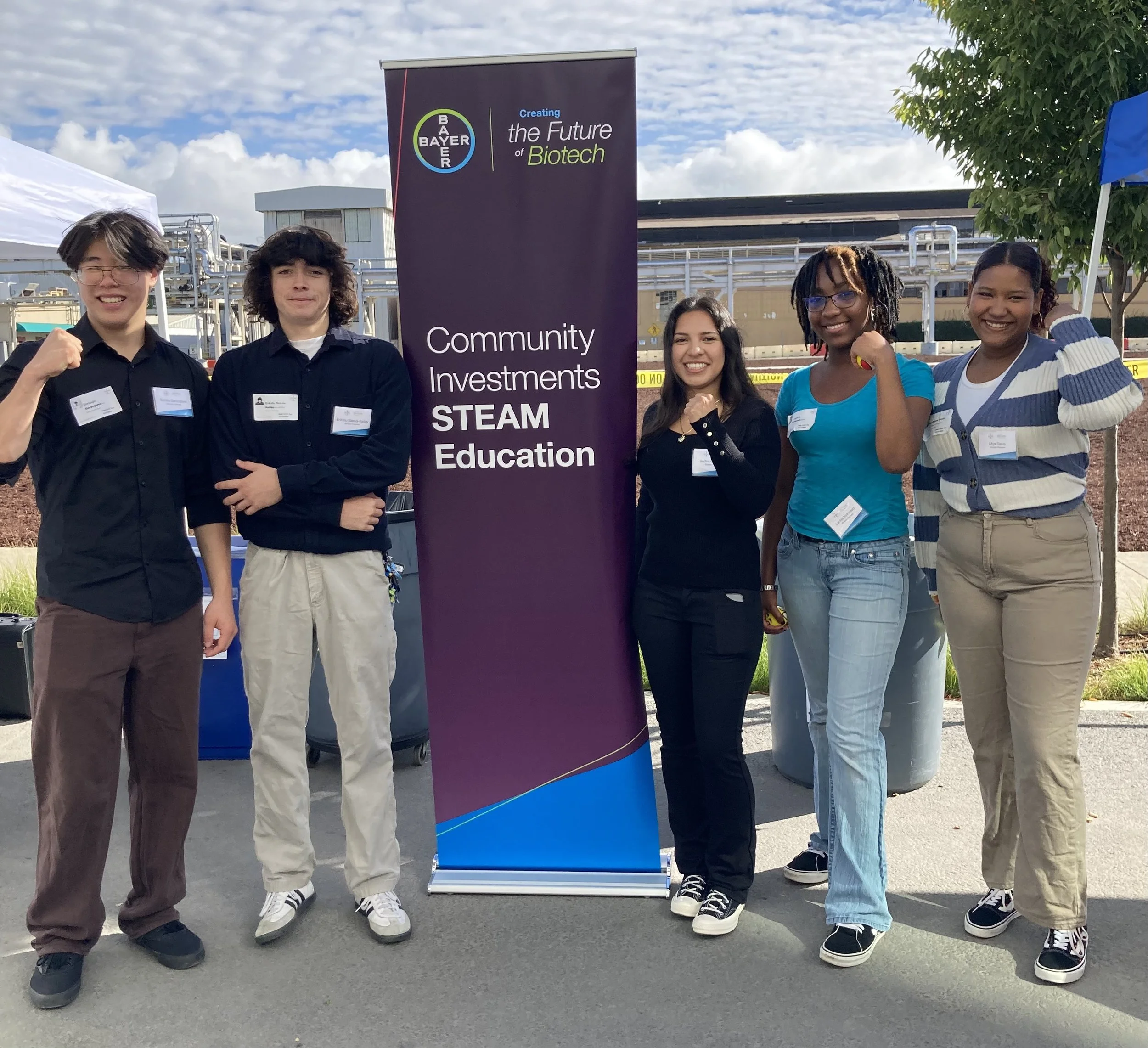 Group of five diverse individuals standing outdoors near a Bayer banner promoting community investments in STEM education, with a cloudy sky and industrial background.