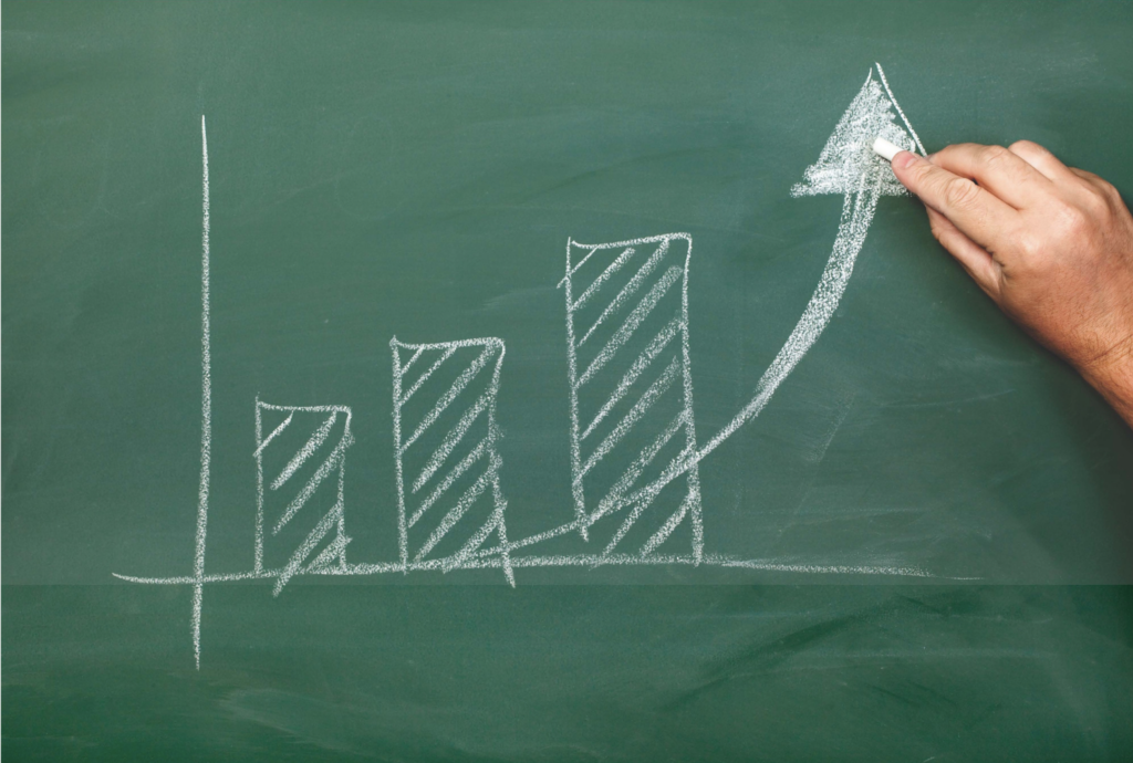 A green chalkboard with a hand drawing a bar graph and an upward arrow with white chalk.