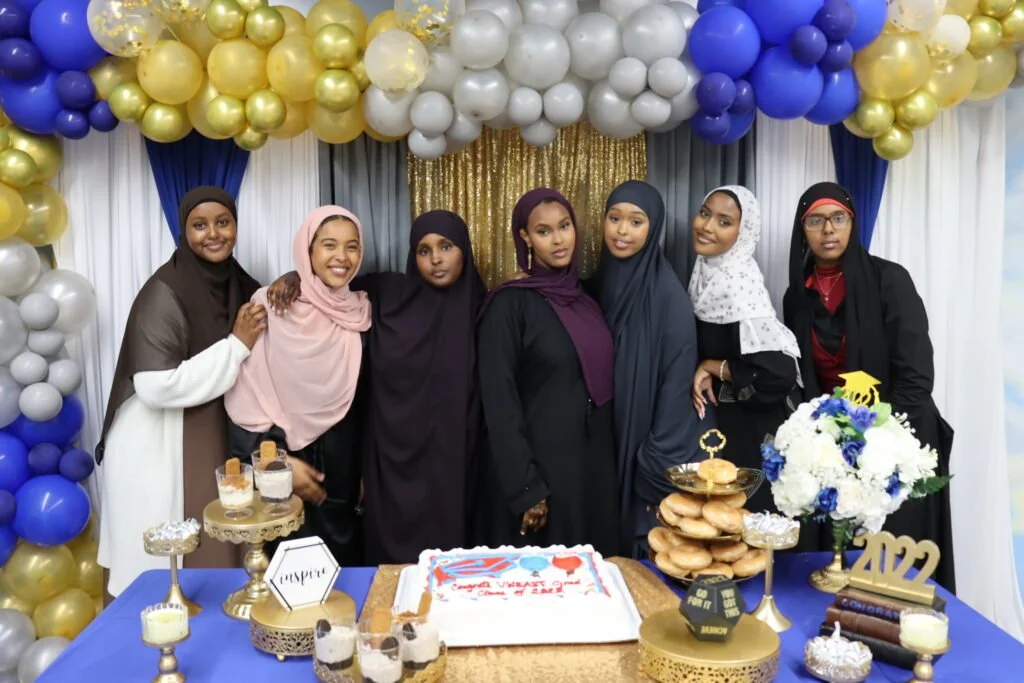 Group of seven women wearing hijabs and dresses at a graduation celebration with decorations, cake, and snacks.