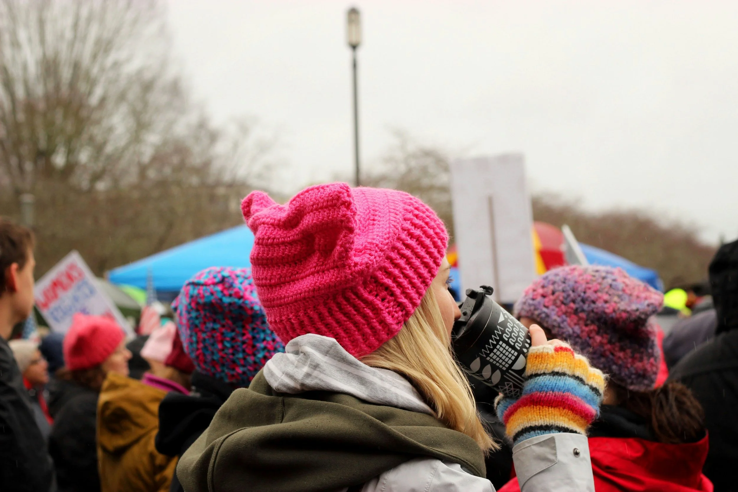 Crowd of people participating in a protest or rally outdoors, many wearing colorful knitted hats and winter clothing, with one woman drinking from a black travel mug.