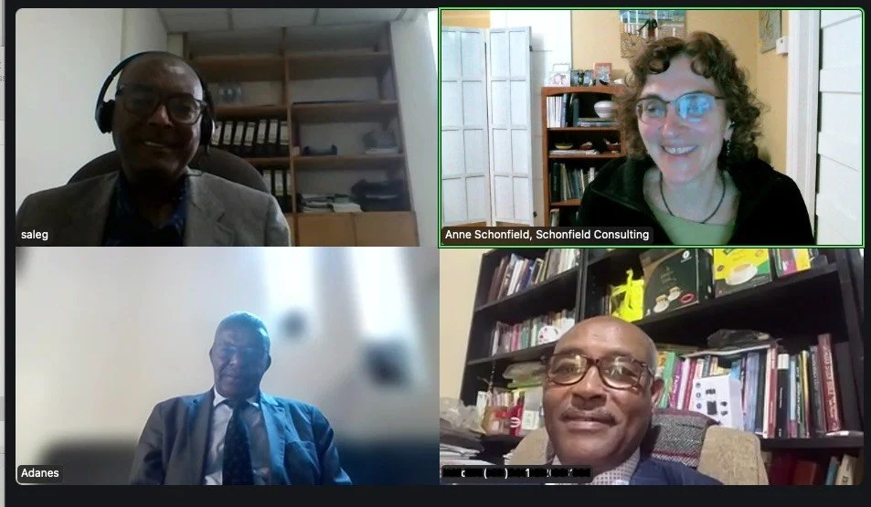 Four people in a virtual video conference, each in their own home office setting. Top left is a man with glasses and headphones, top right is a woman with curly hair and glasses, bottom left is a man in a suit, bottom right is a man with glasses sitting in front of bookshelves.
