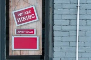 A sign on a brick wall indicates a business is hiring, with messages 'We Are Hiring' and 'Apply Today.'