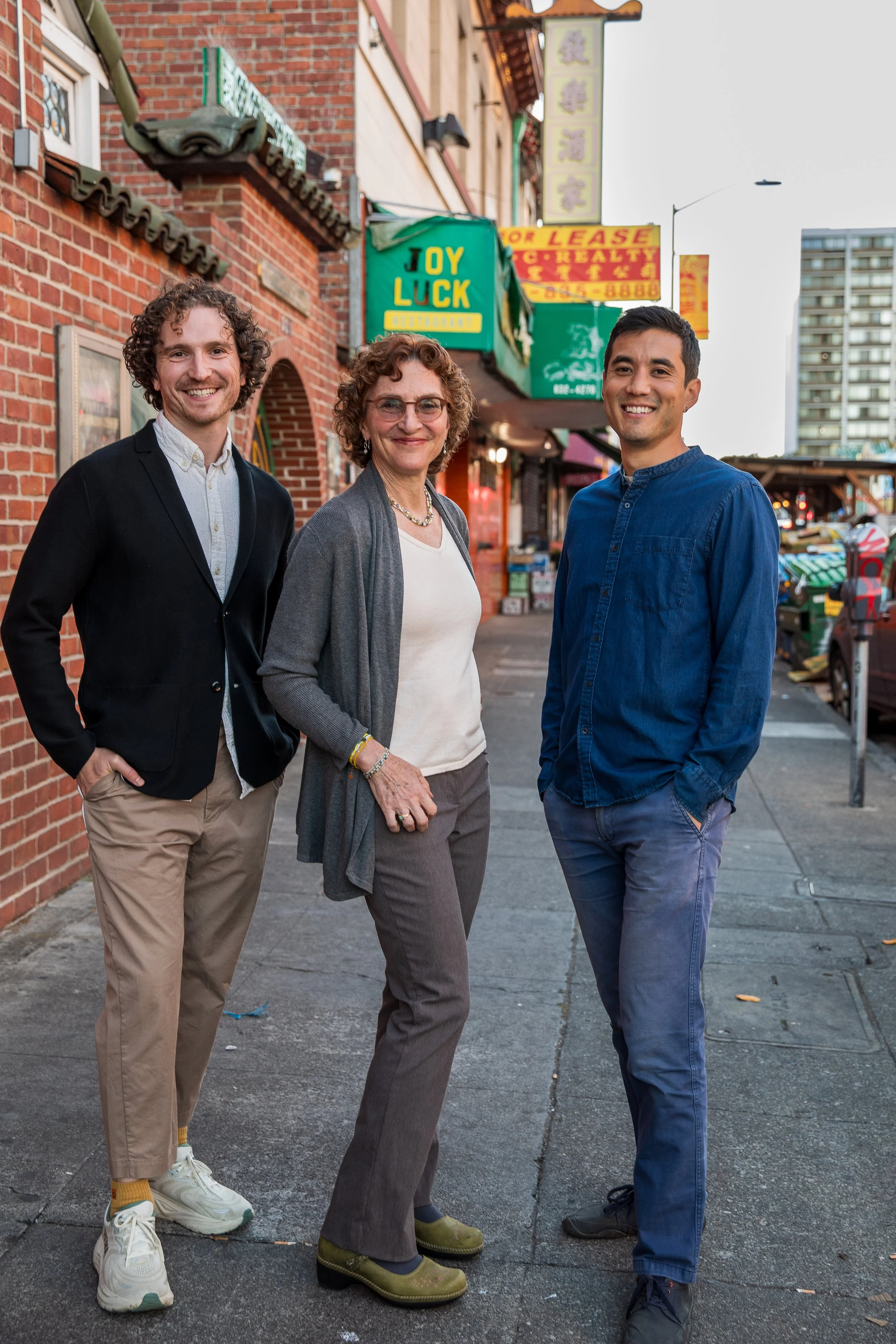 Three people standing on a sidewalk in front of a brick building with a green sign that says 'Joy Luck.' They are smiling and dressed casually.