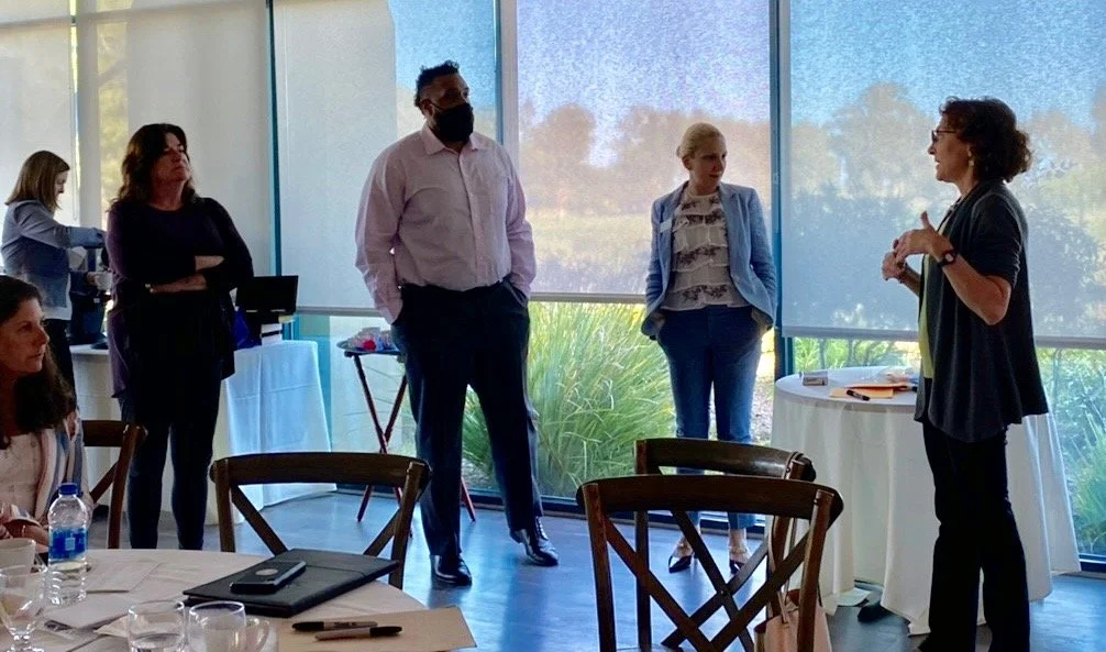 A group of five people socializing in a bright room with large windows at a professional or networking event. Two women are standing near the windows, one talking to the woman on the right, who is wearing glasses and a black blazer. A man in a pink shirt and face mask is standing with his hands in his pockets. Another woman is standing with her arms crossed, and a woman is sitting at a table with water bottles and notebooks.