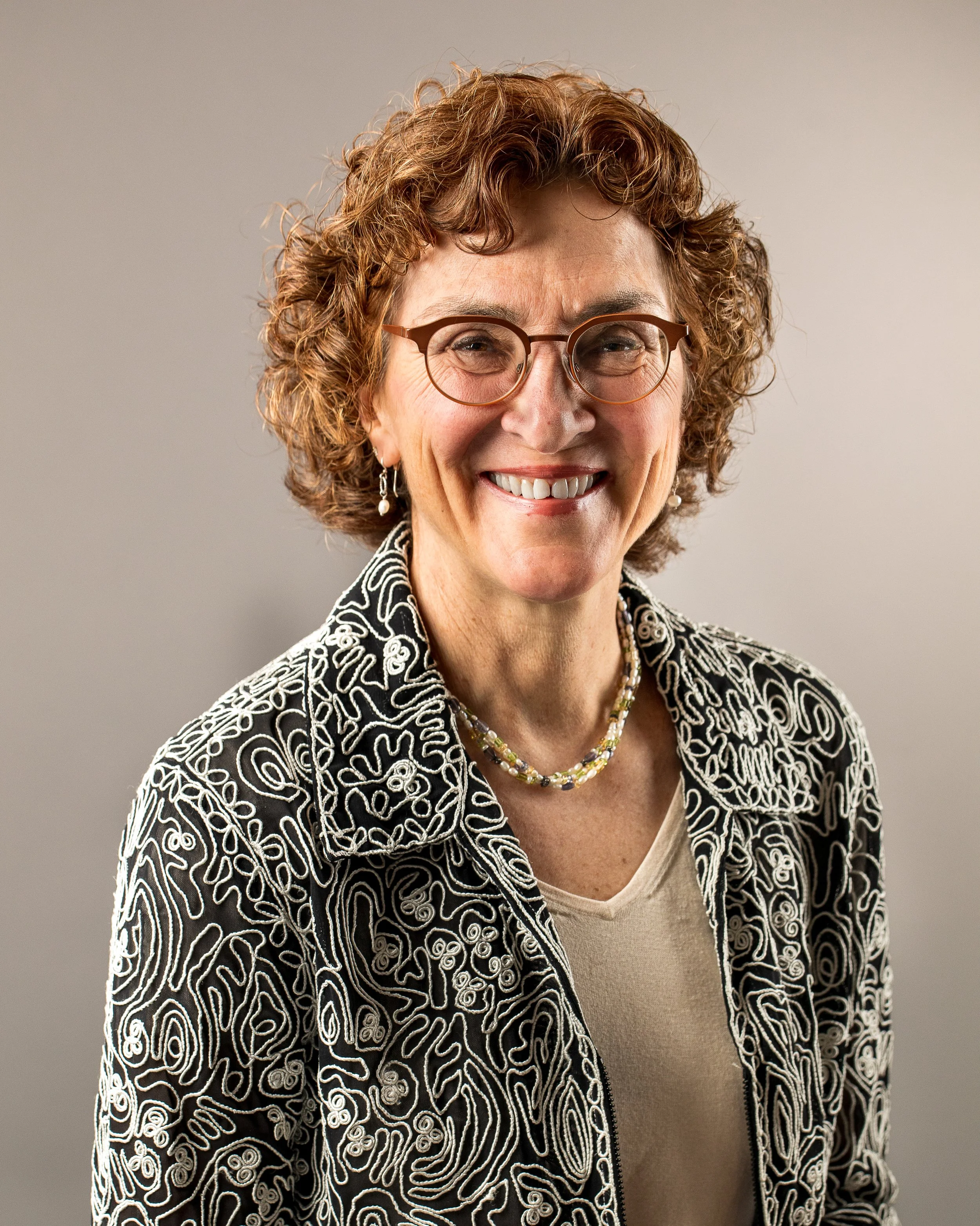 A smiling woman with curly brown hair, glasses, and pearl earrings, wearing a patterned black and white jacket and a beaded necklace, against a plain gray background.
