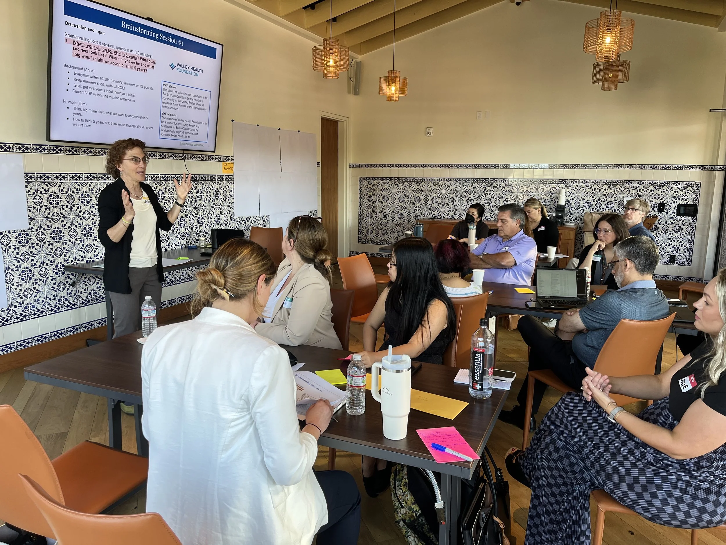 A woman presenting during a brainstorming session in a conference room with an audience of men and women seated at tables. The room features decorative blue and white tile walls, pendant lighting, and a large screen displaying the session details.