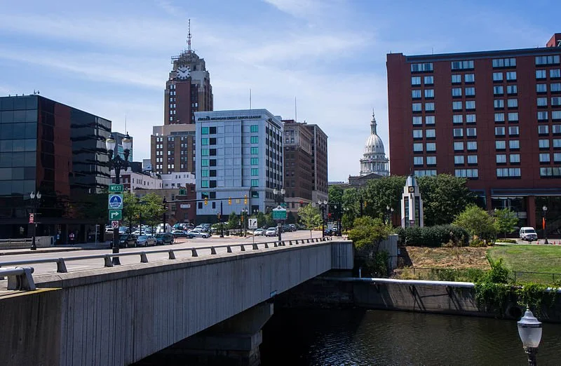 City skyline with tall buildings, a bridge over water, and a government building with a dome in the background.