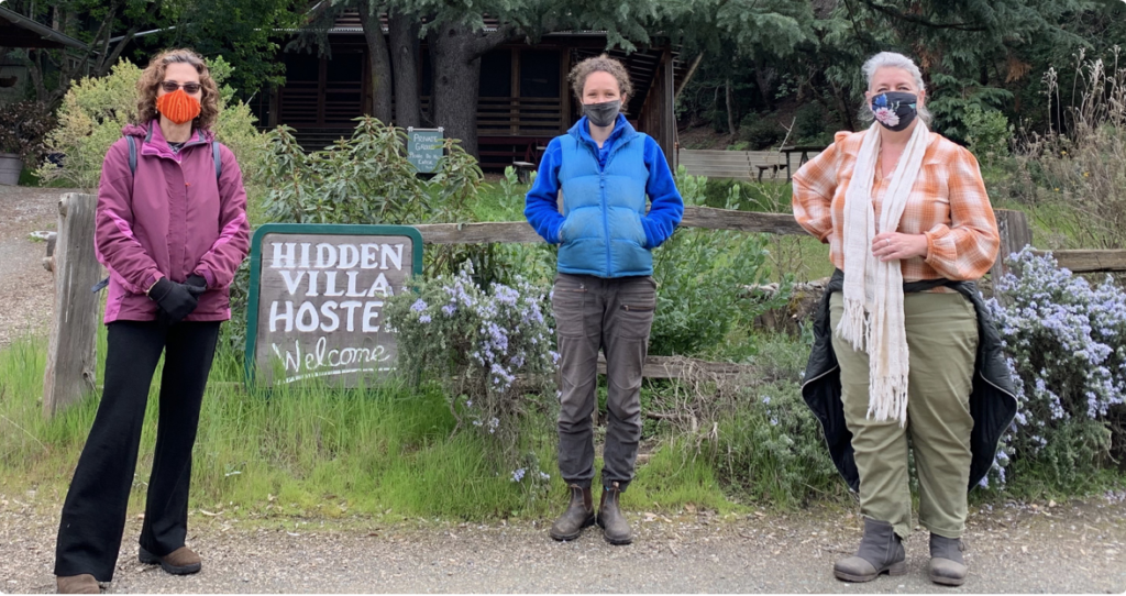 Three people standing outdoors in front of a sign that reads "HIDDEN VILLA HOSTEL Welcome" and some bushes and trees, all wearing masks.