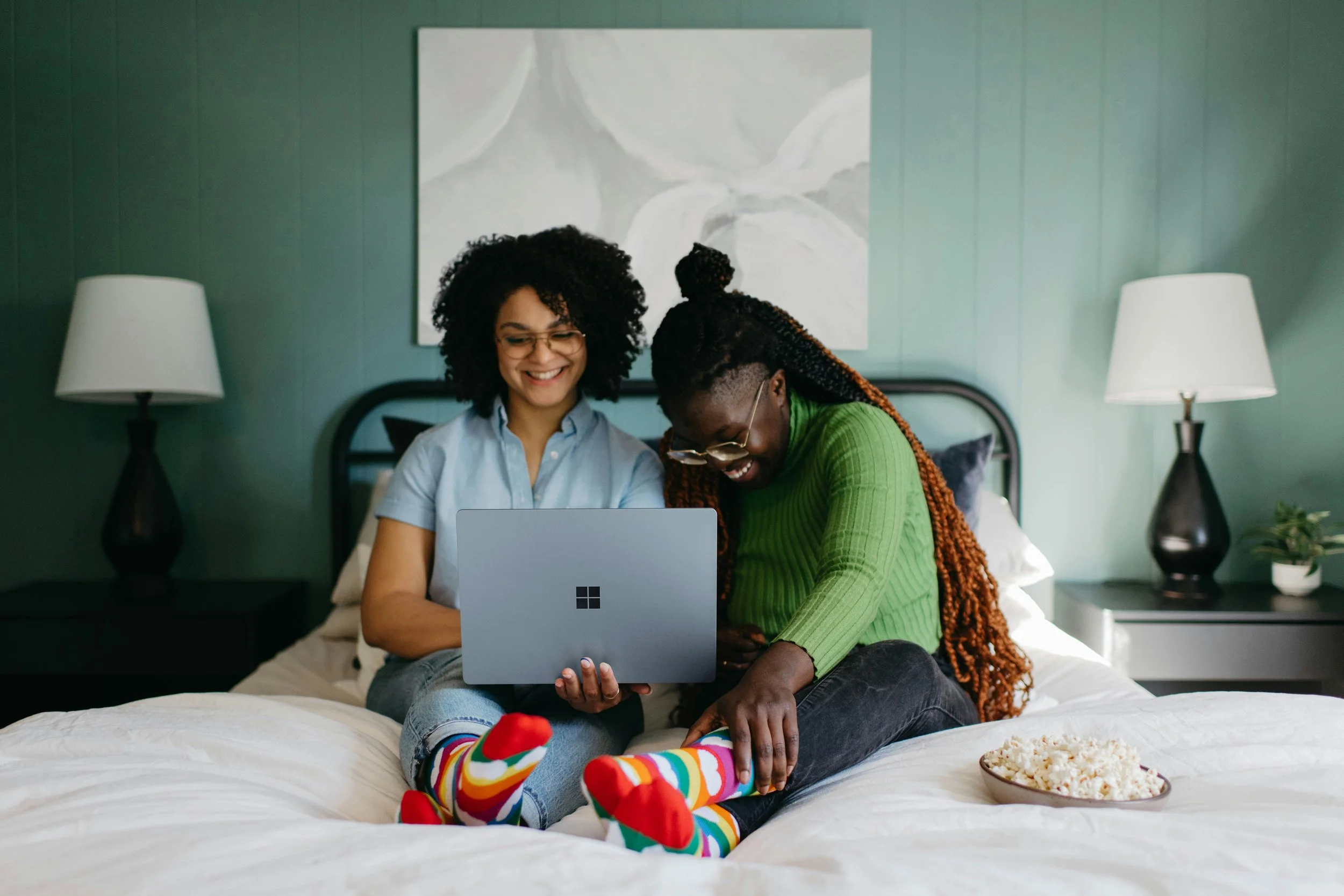 Two women sitting on a bed, smiling and looking at a laptop, in a bedroom with green wallpaper and two bedside lamps, one on each side. One woman is wearing glasses and a light blue shirt, while the other has dreadlocks and is wearing a green sweater. A bowl of popcorn is on the bed beside them.