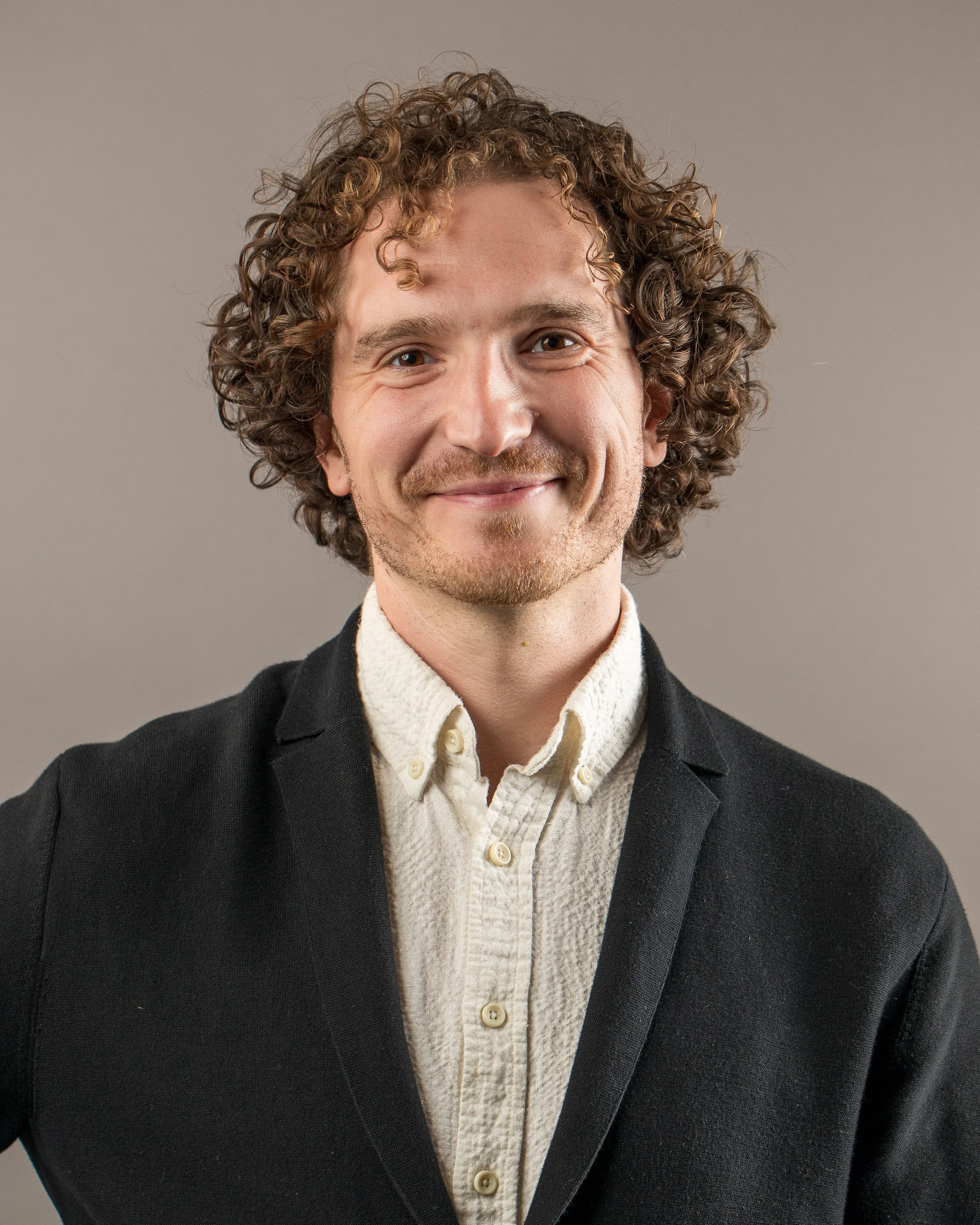 A man with curly hair and a light beard wearing a beige shirt and black blazer, smiling at the camera against a plain gray background.
