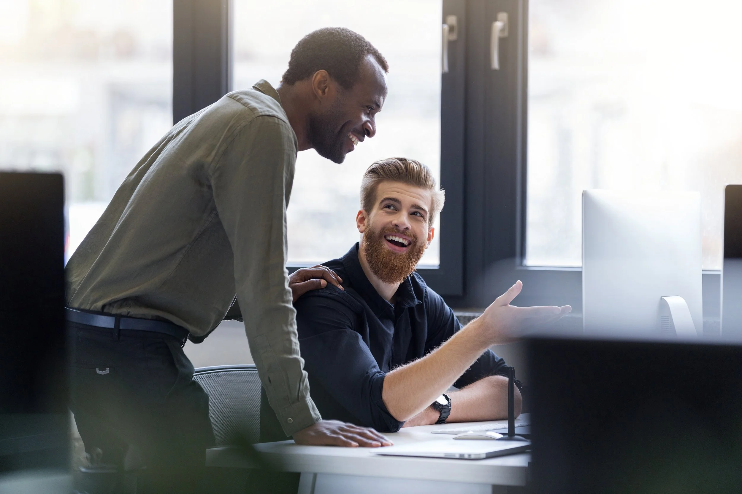 Two men in an office, smiling and having a conversation near a computer, with large windows in the background.