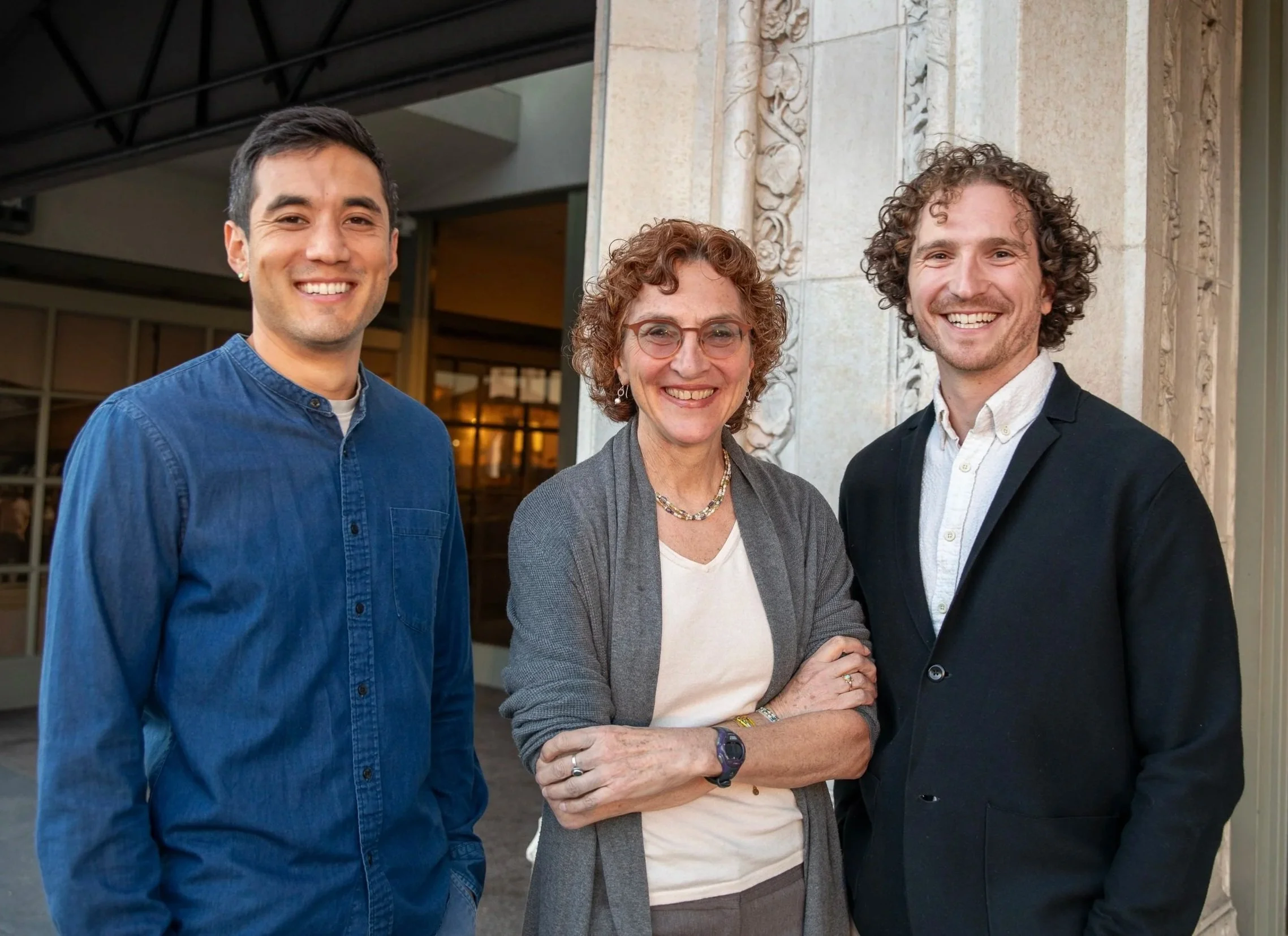 Three smiling people standing together outdoors in front of a building with decorative stonework. The group includes a young man in a blue shirt, a woman with glasses and curly hair in a gray blazer, and another young man in a black blazer with curly hair.
