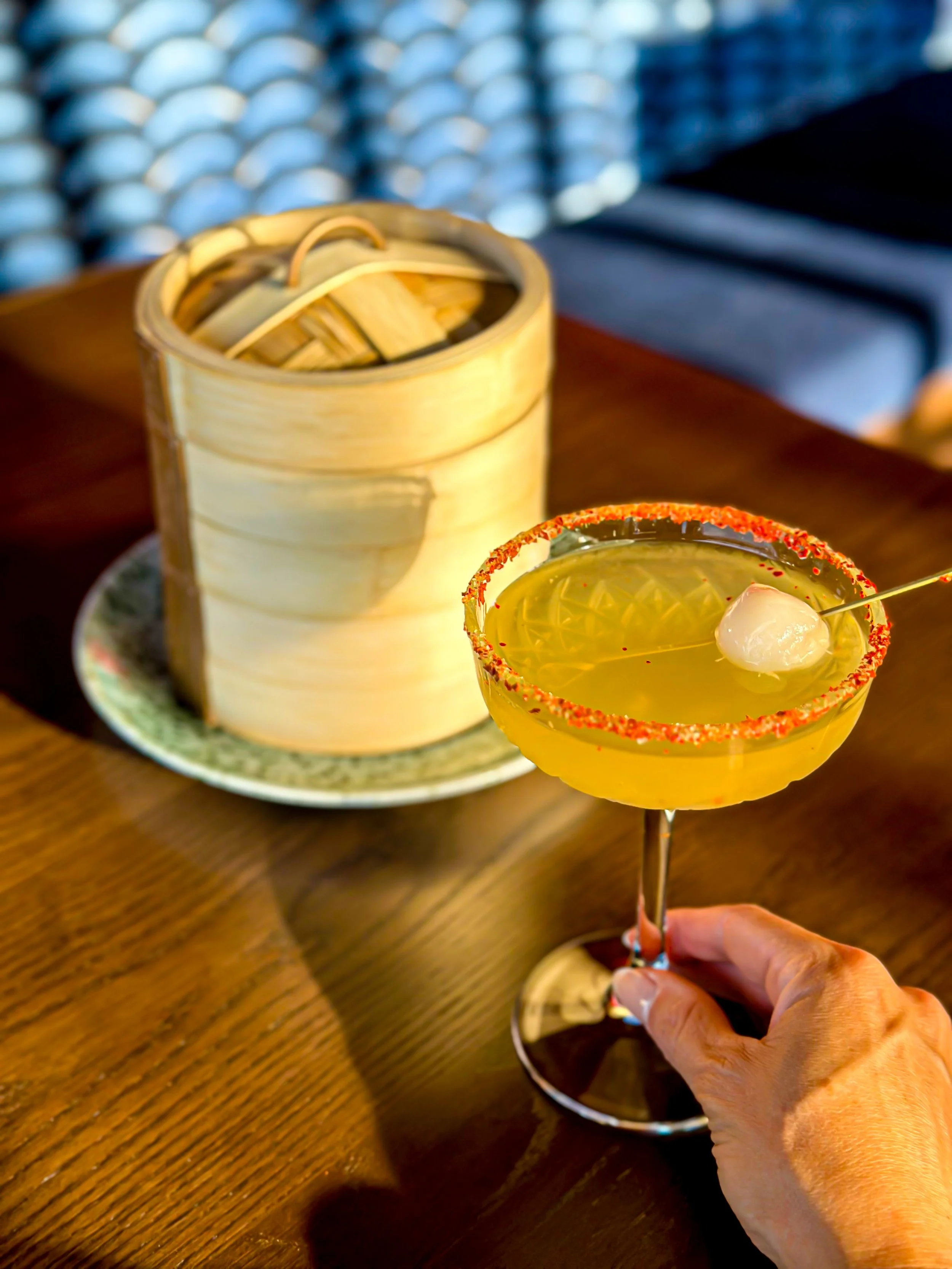 A cocktail in a margarita glass with a salted rim and a small skewer of fruit inside, held by a hand. In the background, a bamboo steamer is placed on a plate on a wooden table.