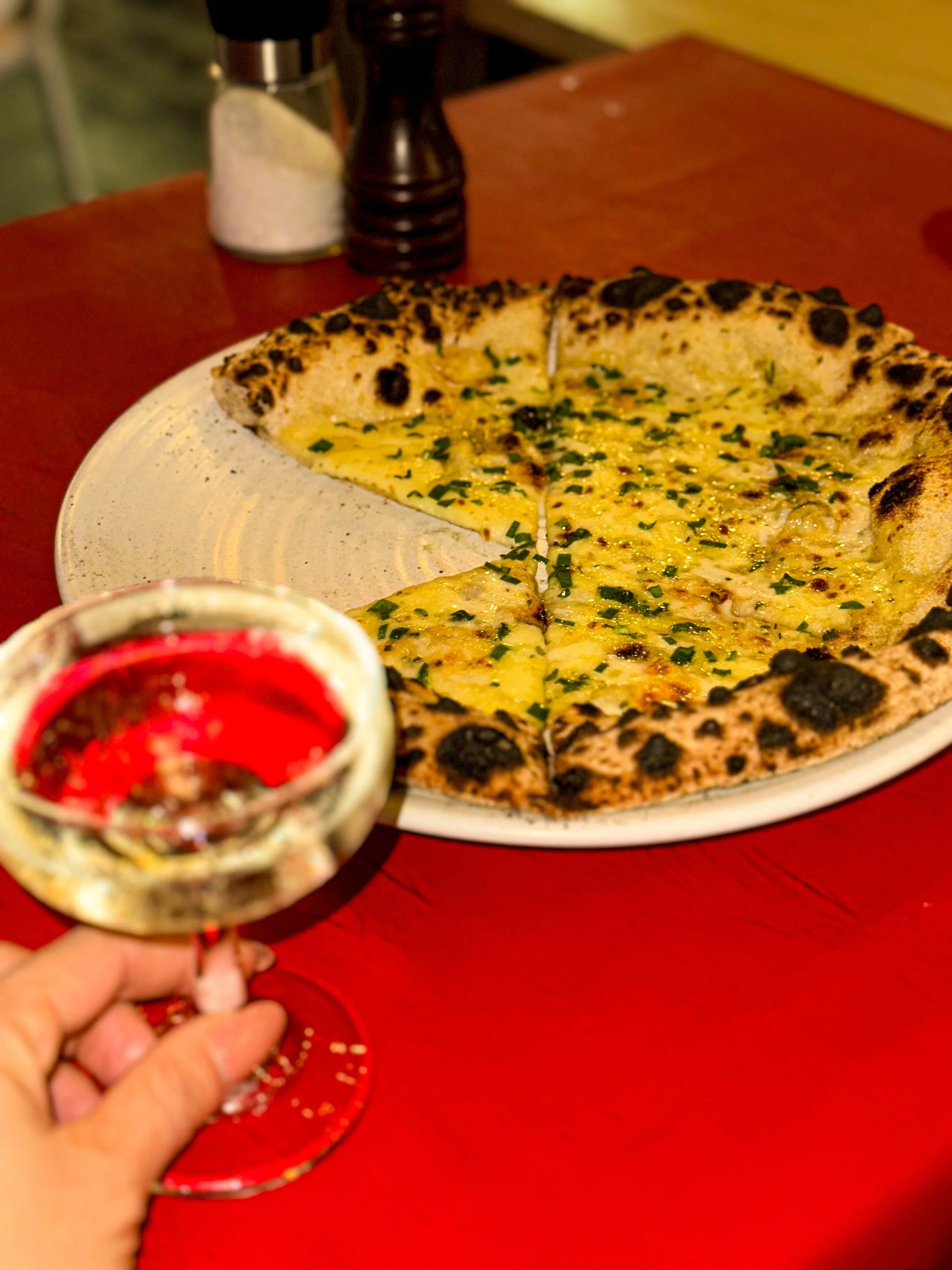 A wood-fired pizza topped with cheese and chopped herbs on a white plate, with a glass of red wine in the foreground. Salt and pepper shakers are in the background on a red table.