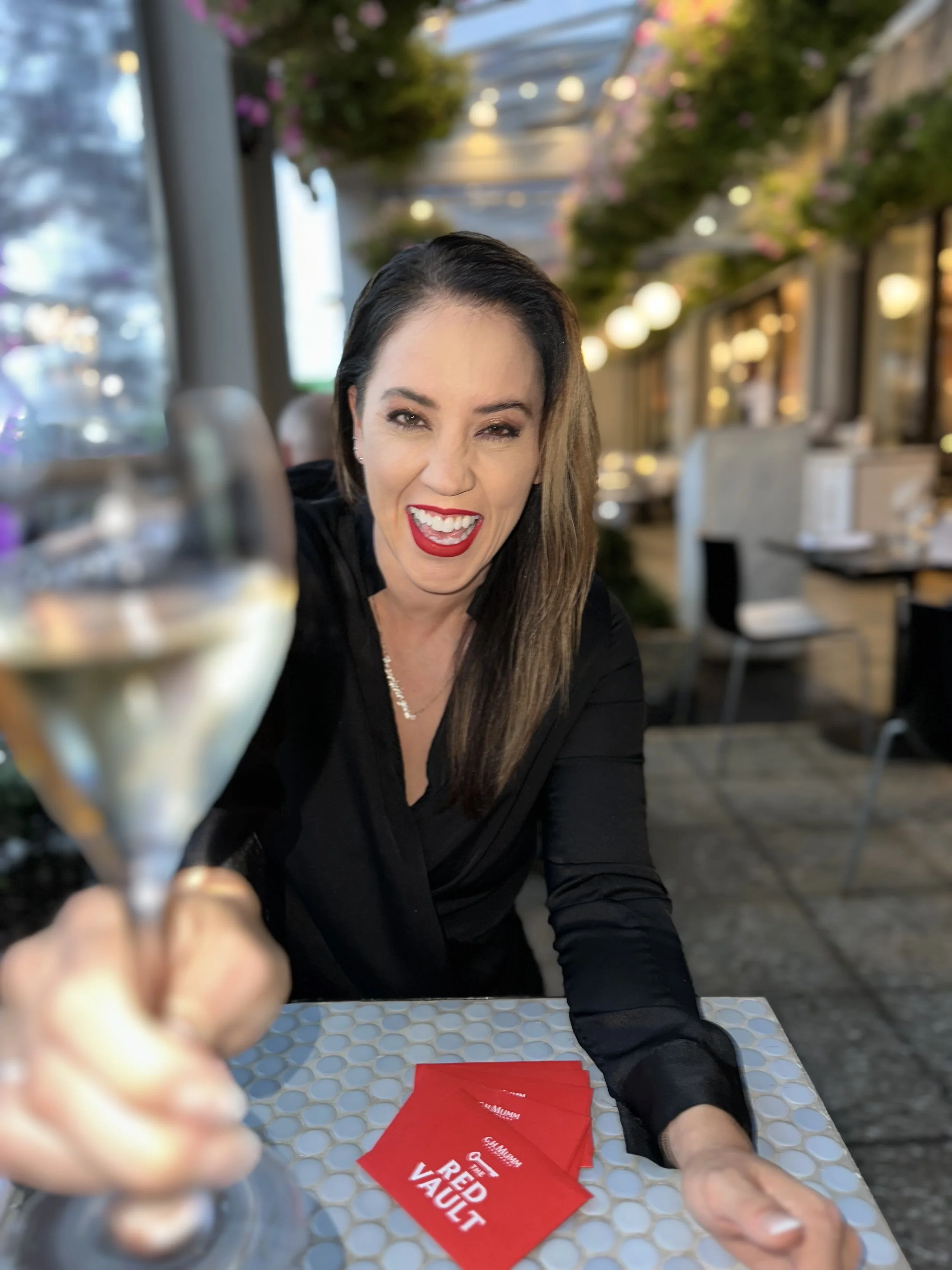 Chan Can Eat. Woman with long dark hair and red lipstick cheers with a glass of white wine at an outdoor restaurant, smiling and winking at the camera.