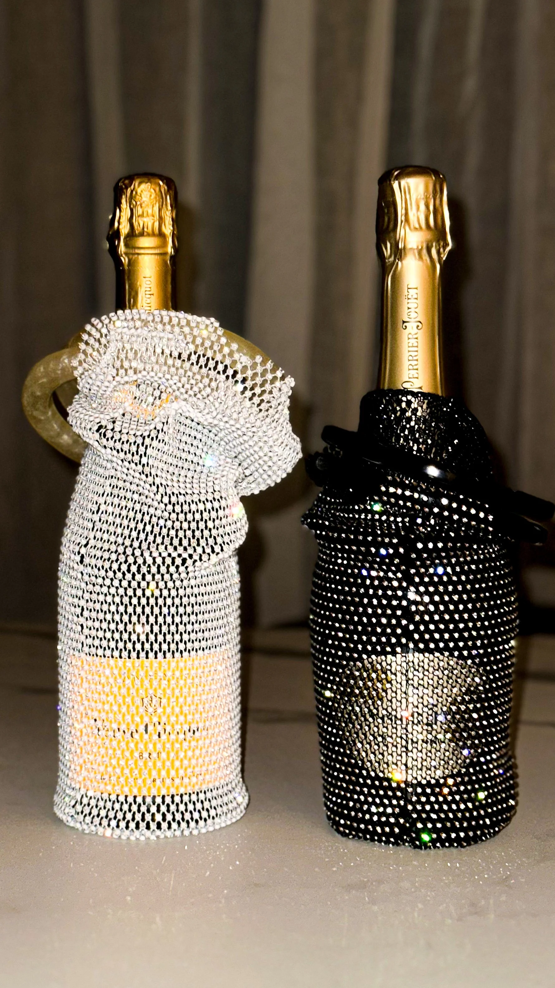 Two champagne bottles in black and white diamond-studded holders with a note on the bottles reading 'PERFIER JOUET' and 'Louis Vuitton' with a gray curtain background.