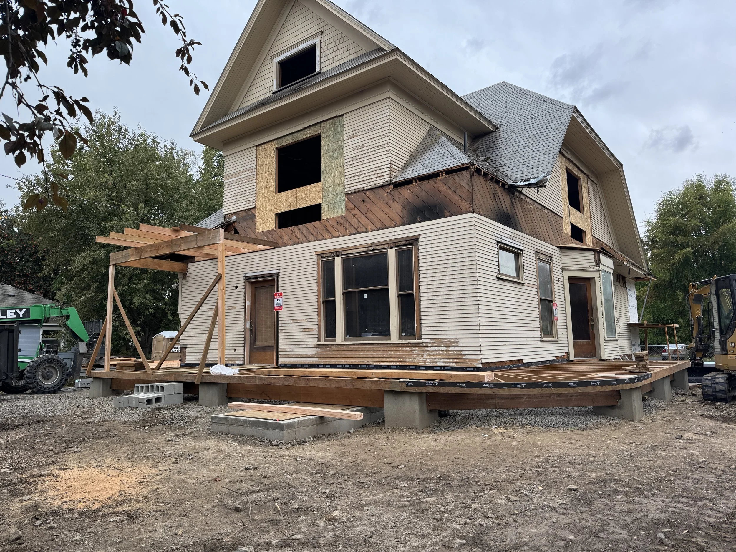 A house under construction with weathered siding partially covered by new plywood, elevated on concrete blocks with a wooden deck foundation, surrounded by construction equipment and trees.