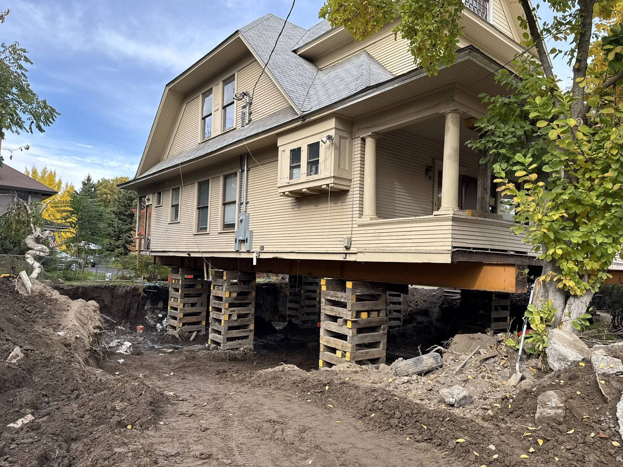 A house undergoing foundation repair with the house elevated on wooden blocks and support beams, and the dirt around the area excavated.