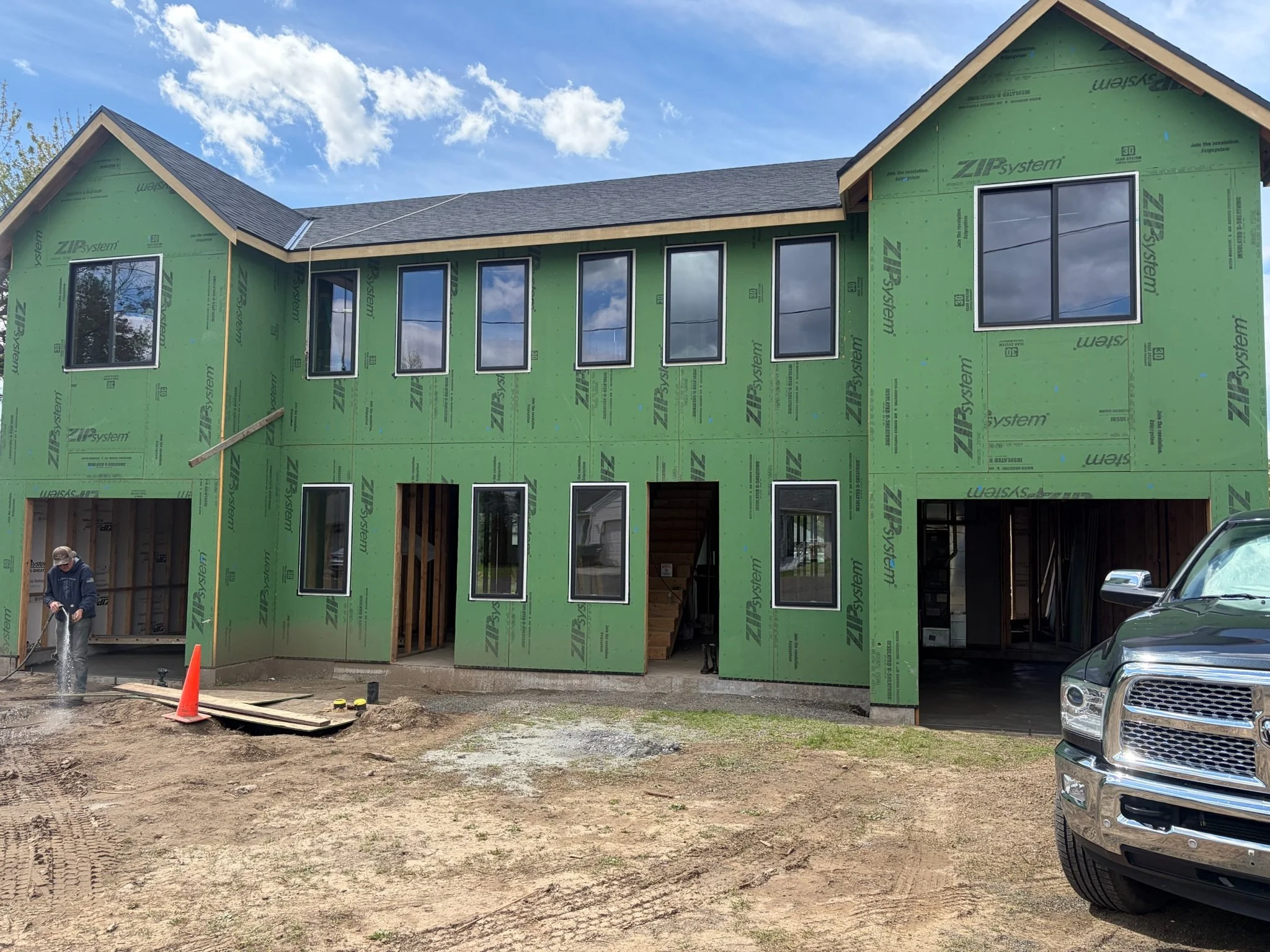 Construction site with a partially built house covered in green sheathing and multiple windows, parked cars, and a worker in a hard hat.