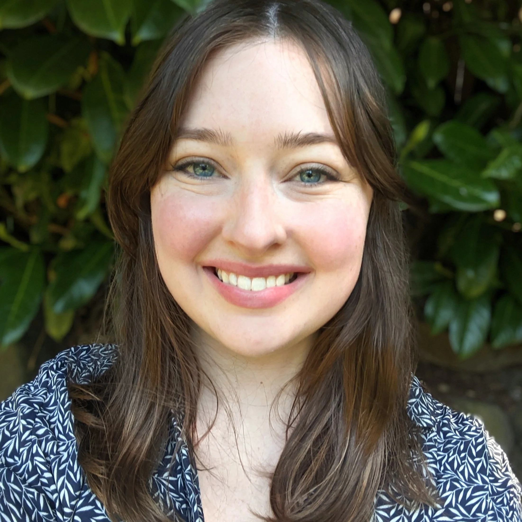 Smiling white woman with brown hair and a navy blue floral shirt in front of foliage