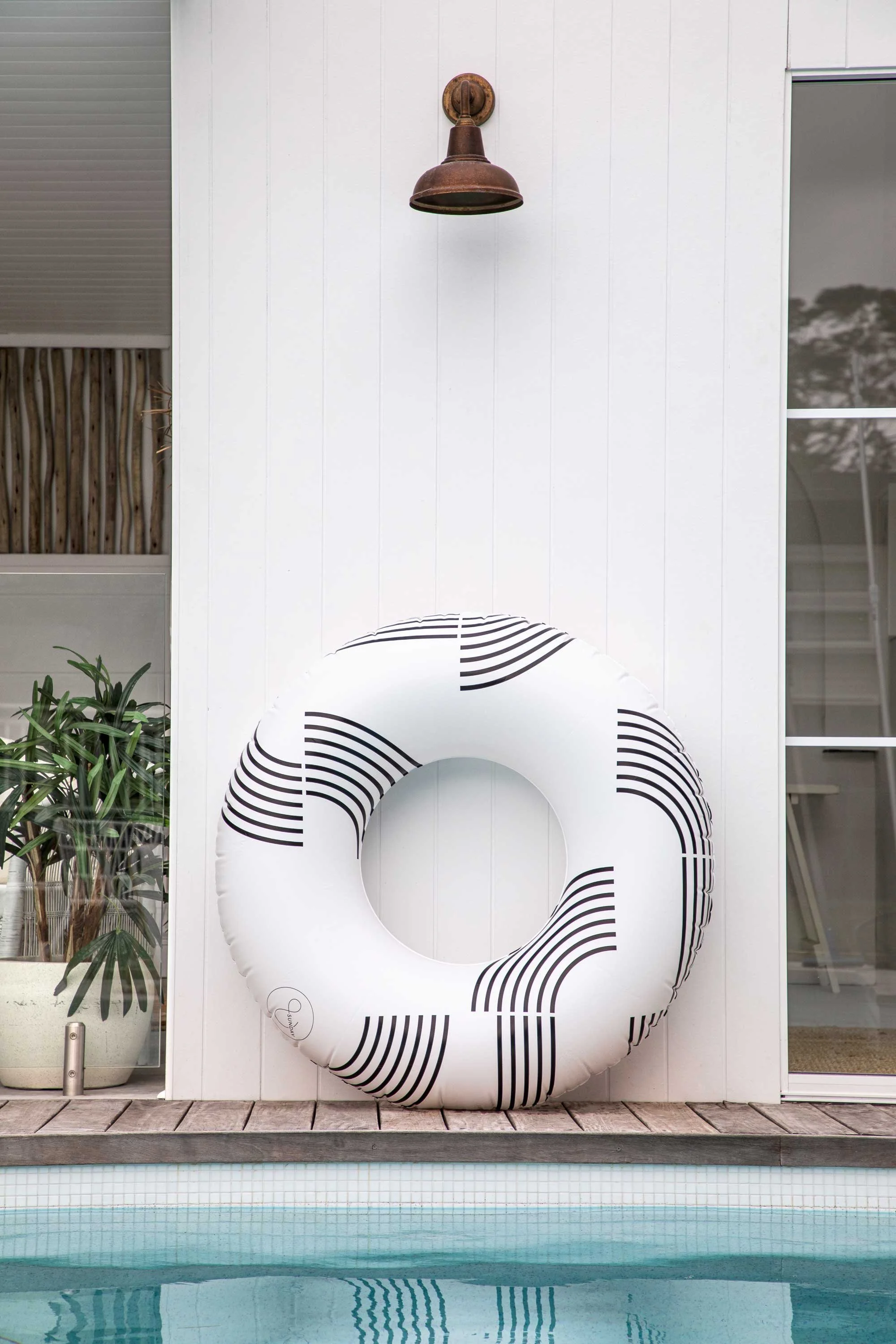 A white wall with a rusted outdoor light fixture above a black and white inflatable pool donut. To the left, a plant in a pot and a window with blinds. In the foreground, part of a swimming pool with water reflecting the white wall and the pool donut