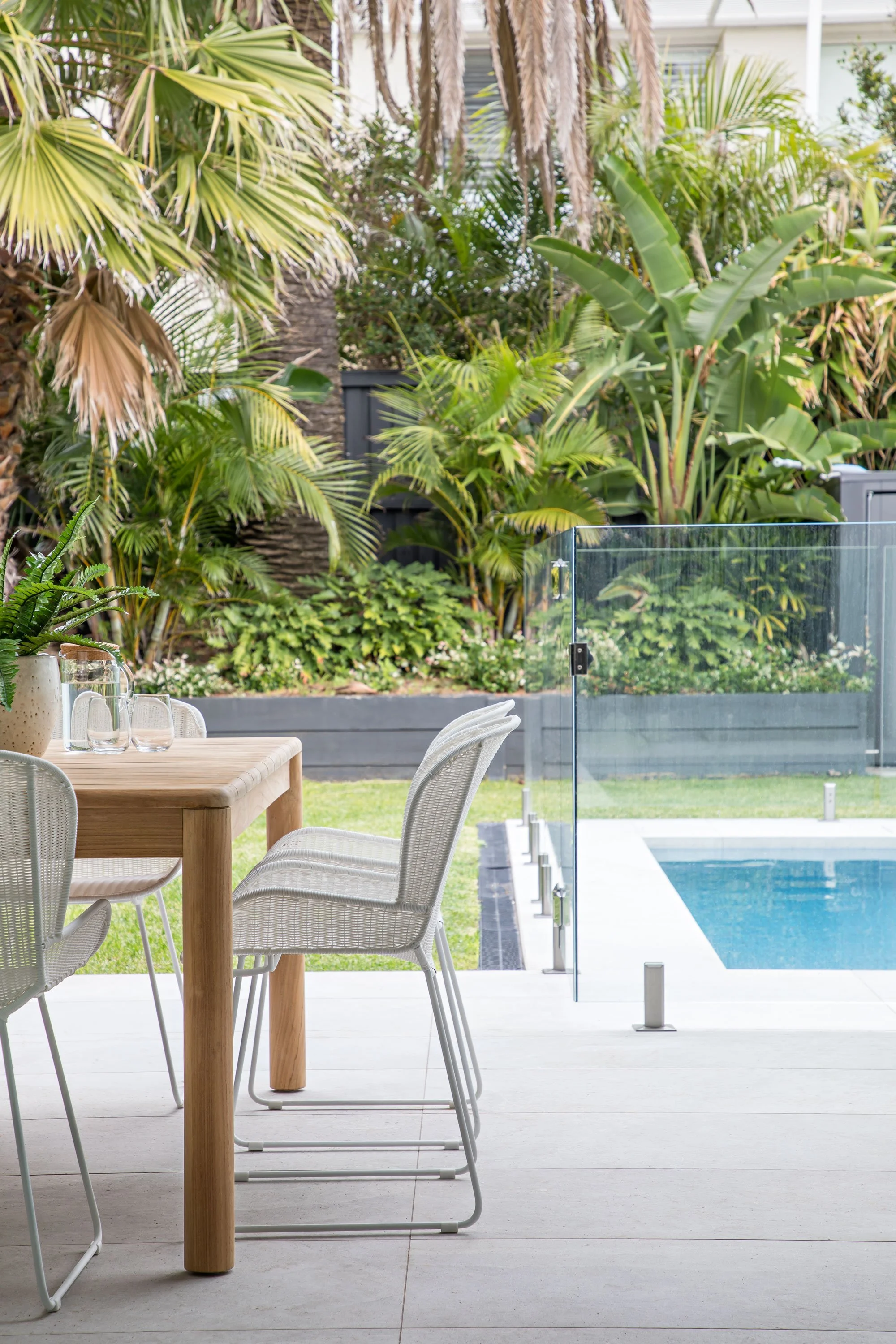 A modern outdoor patio area with a wooden table and white chairs, overlooking a swimming pool, surrounded by lush green tropical plants and trees.