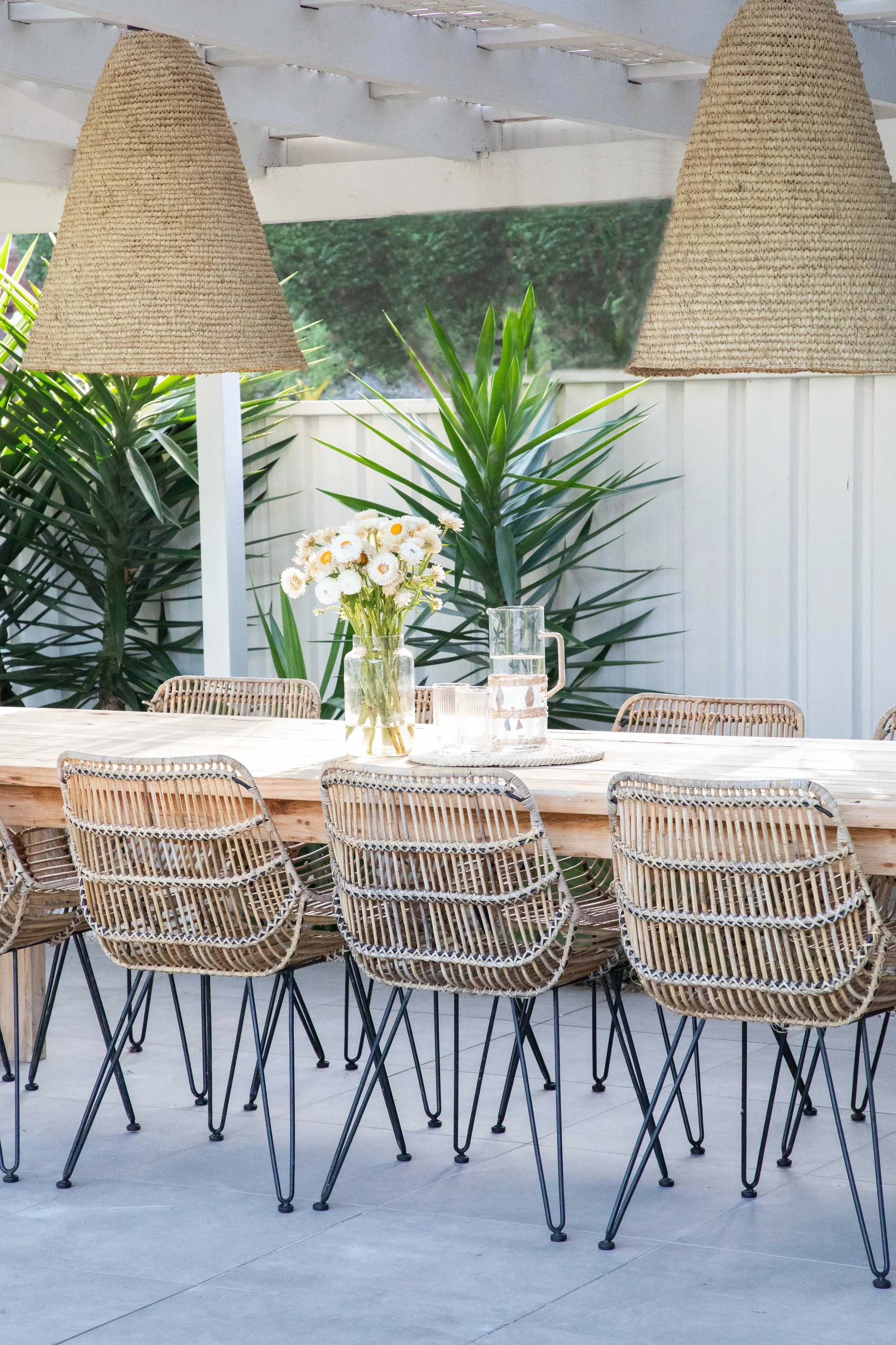 Outdoor dining space with a long wooden table surrounded by woven rattan chairs, decorated with a vase of white daisies, and two hanging pendant lights with woven shades, with lush green plants in the background.