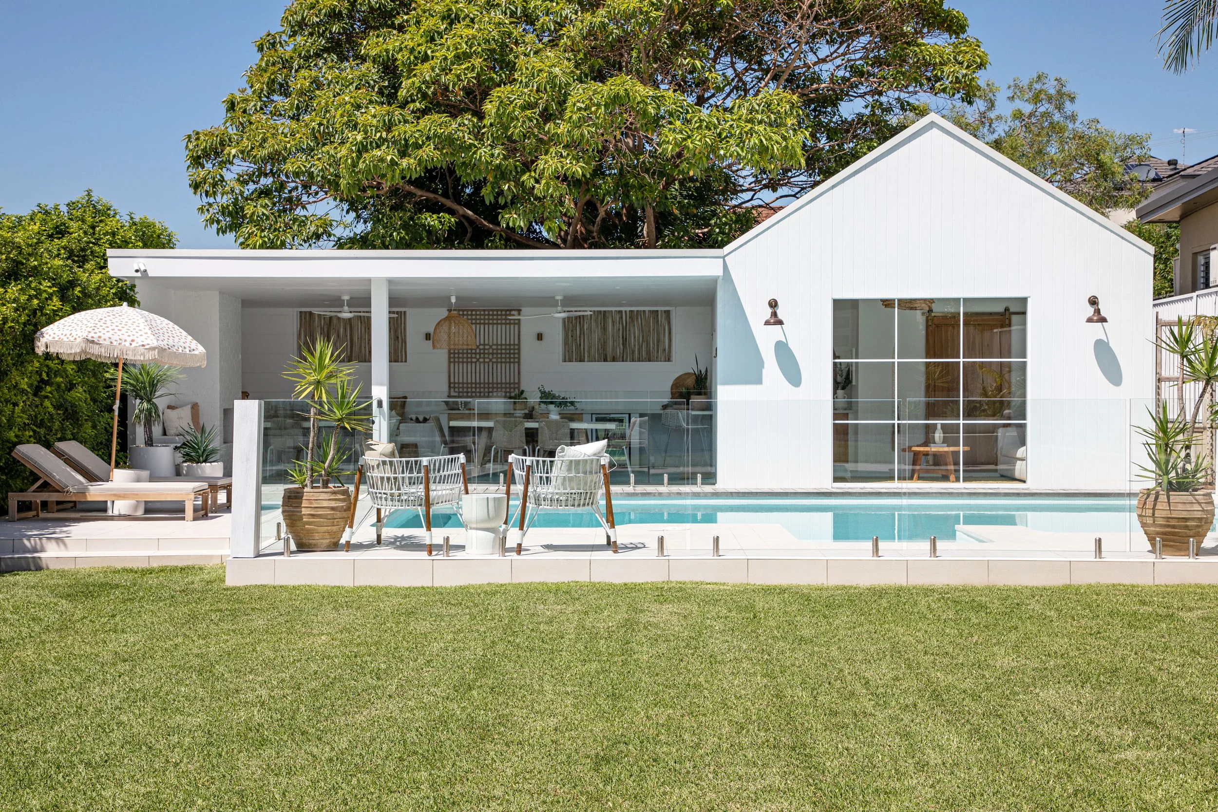 Modern white house with large glass windows and sliding doors, outdoor patio with seating, potted plants, and a swimming pool, surrounded by green lawn and trees under a clear blue sky.