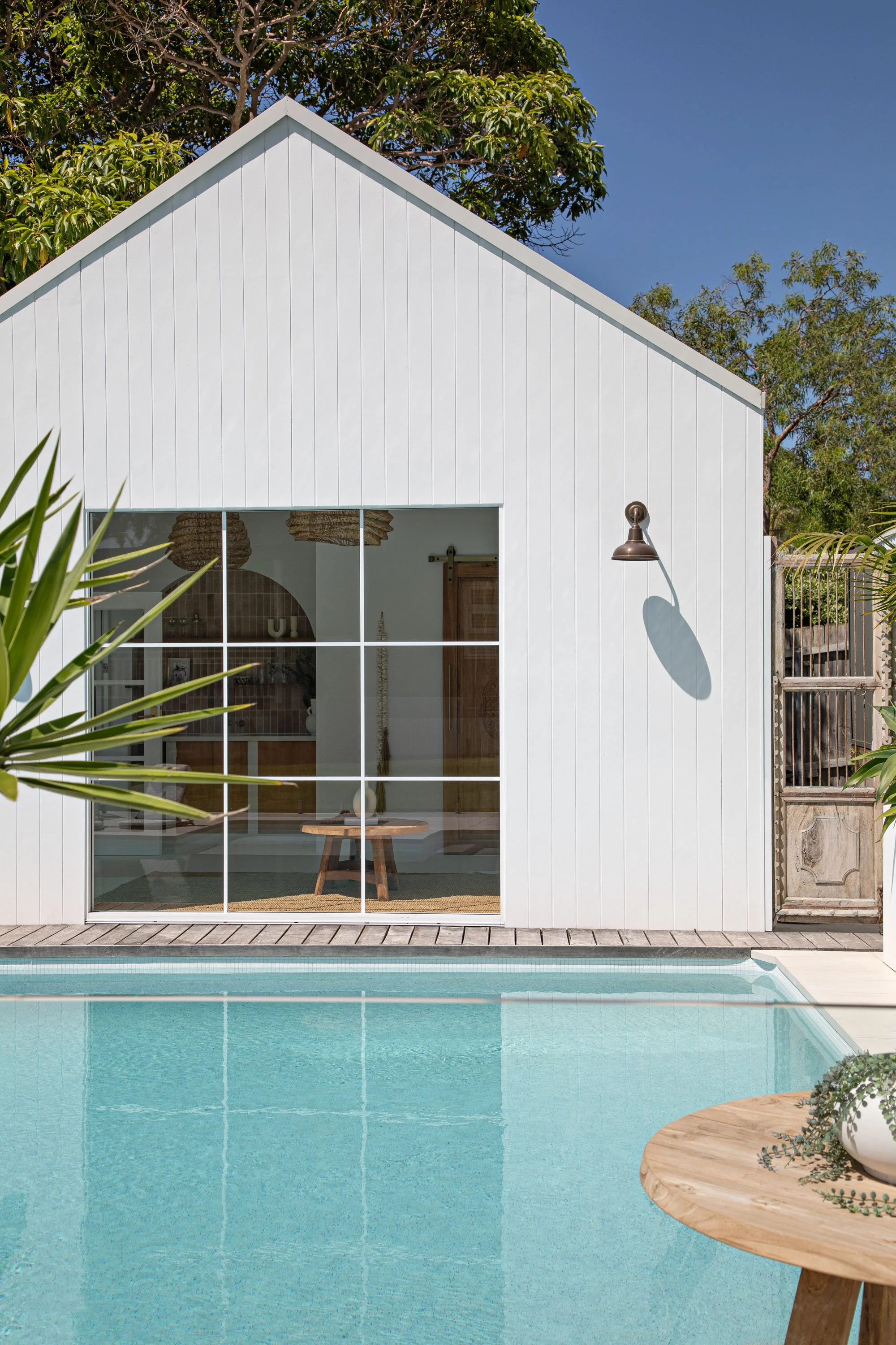 View of a modern house with a large glass wall facing a swimming pool. The house has white vertical siding and a triangular roof. There are trees in the background and a wooden table with a plant on it in the foreground.