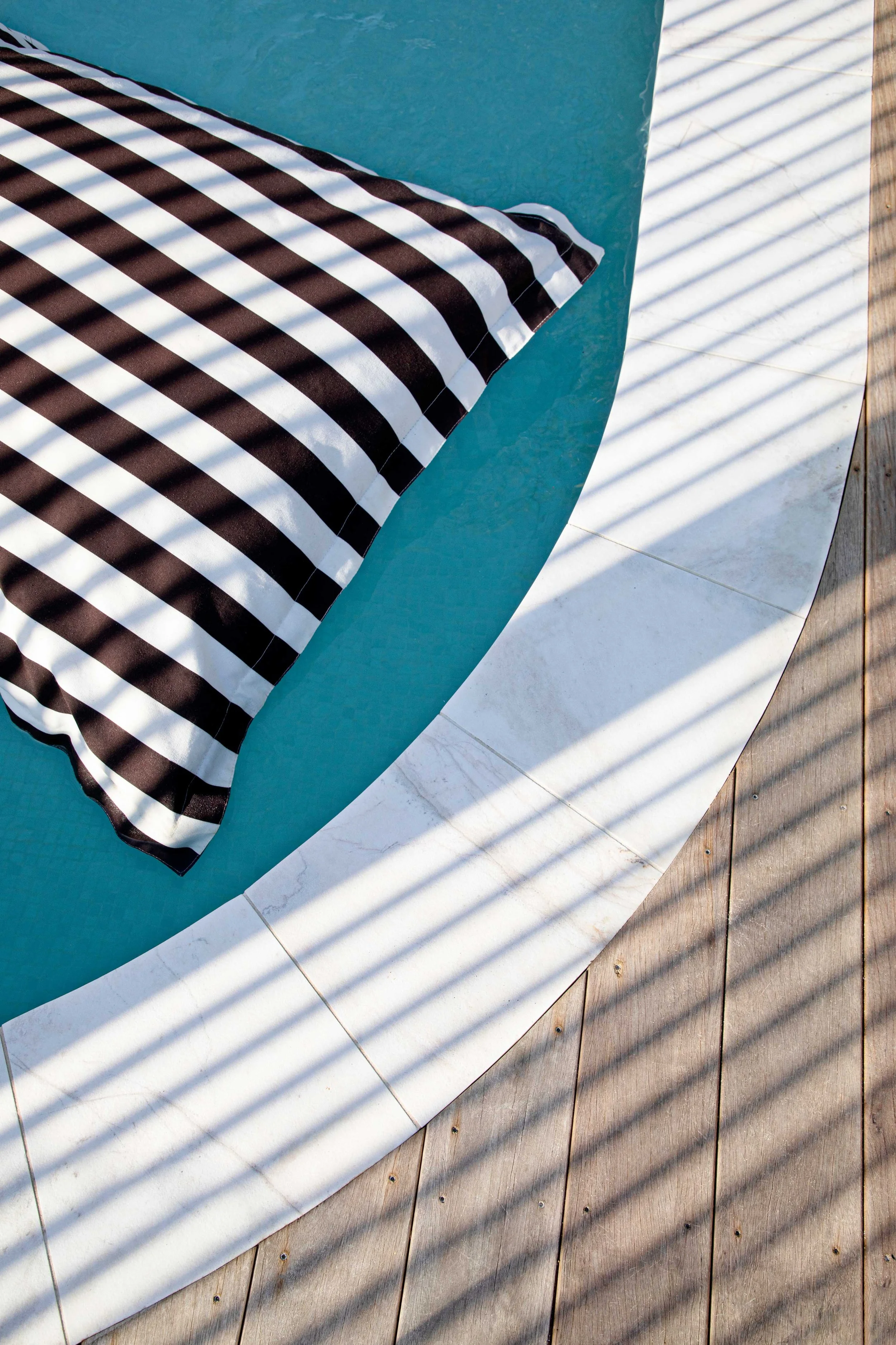 A striped pool pillow floats on a turquoise pool, with shadows cast by a barrier creating a pattern across the scene and wooden deck tiles nearby.