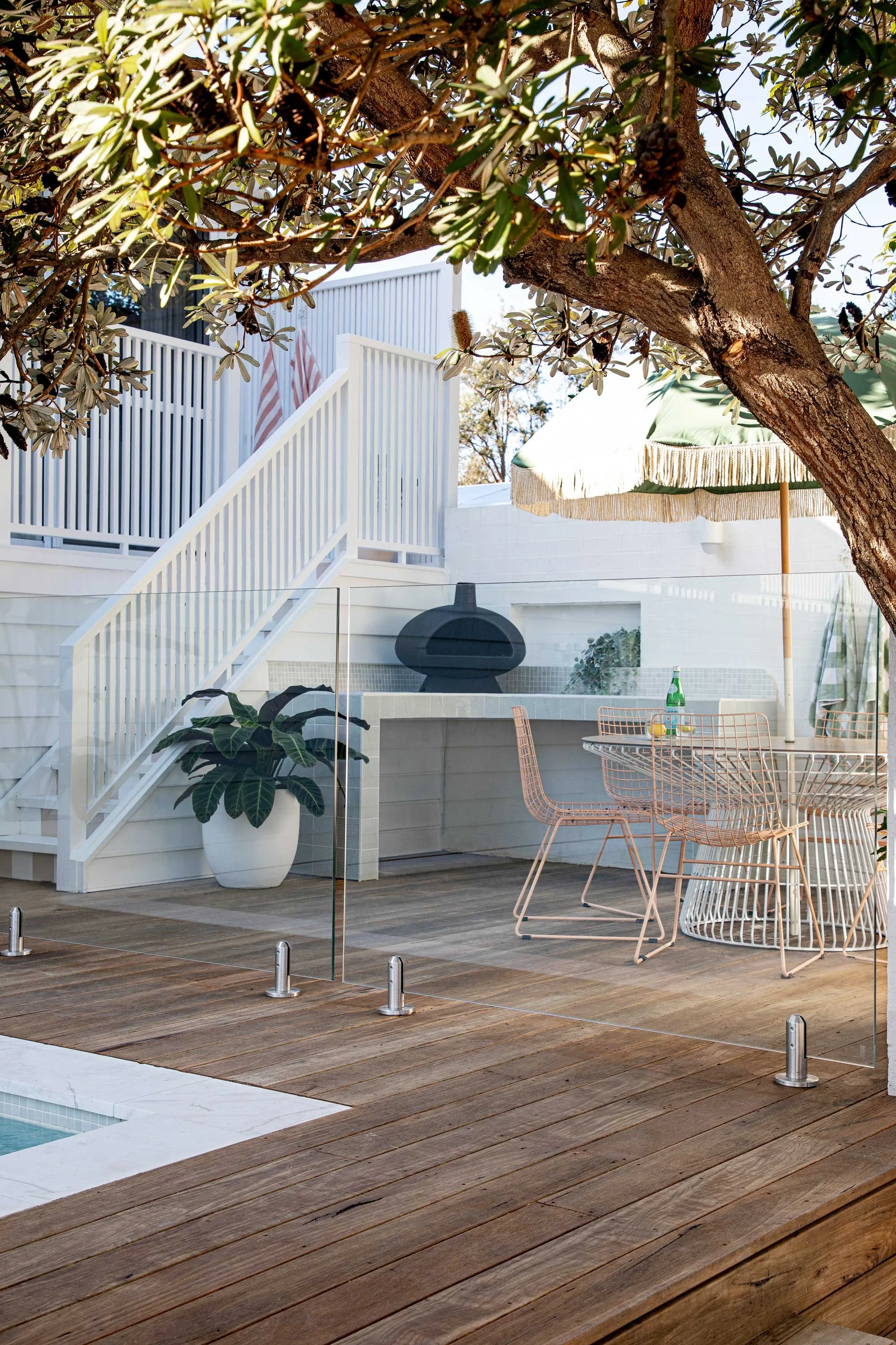 A wooden outdoor deck with a glass panel fence around it, a large potted plant, a black pizza oven, and a round outdoor dining table with pink wire chairs. There is an umbrella providing shade and a tree with dense leaves growing over the deck.