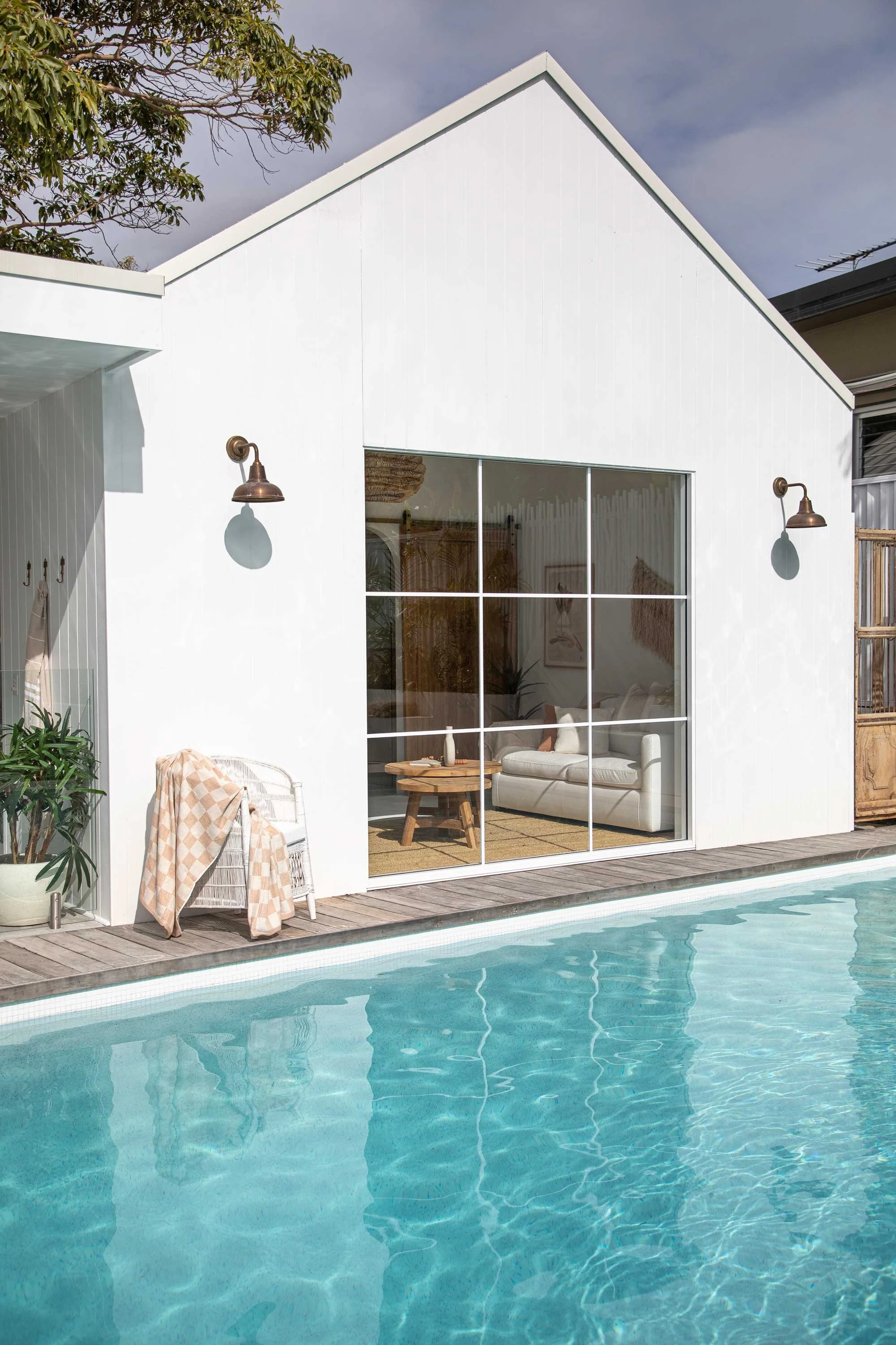 View of a modern white house with large glass sliding doors, adjacent to a swimming pool, with outdoor seating and greenery.
