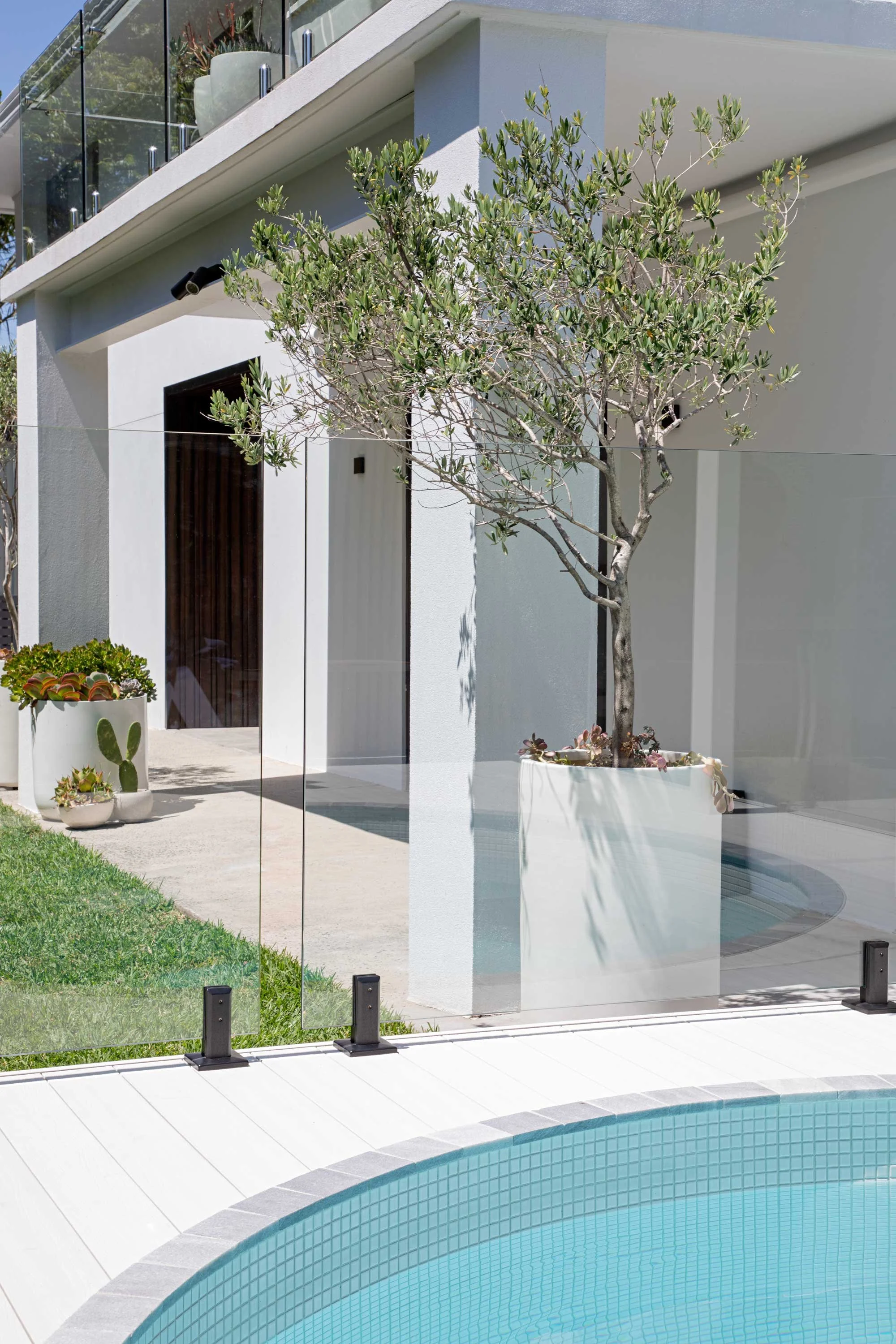 Modern house with white exterior, glass balcony, and small tree in a white planter outside near a swimming pool with tiled lining.