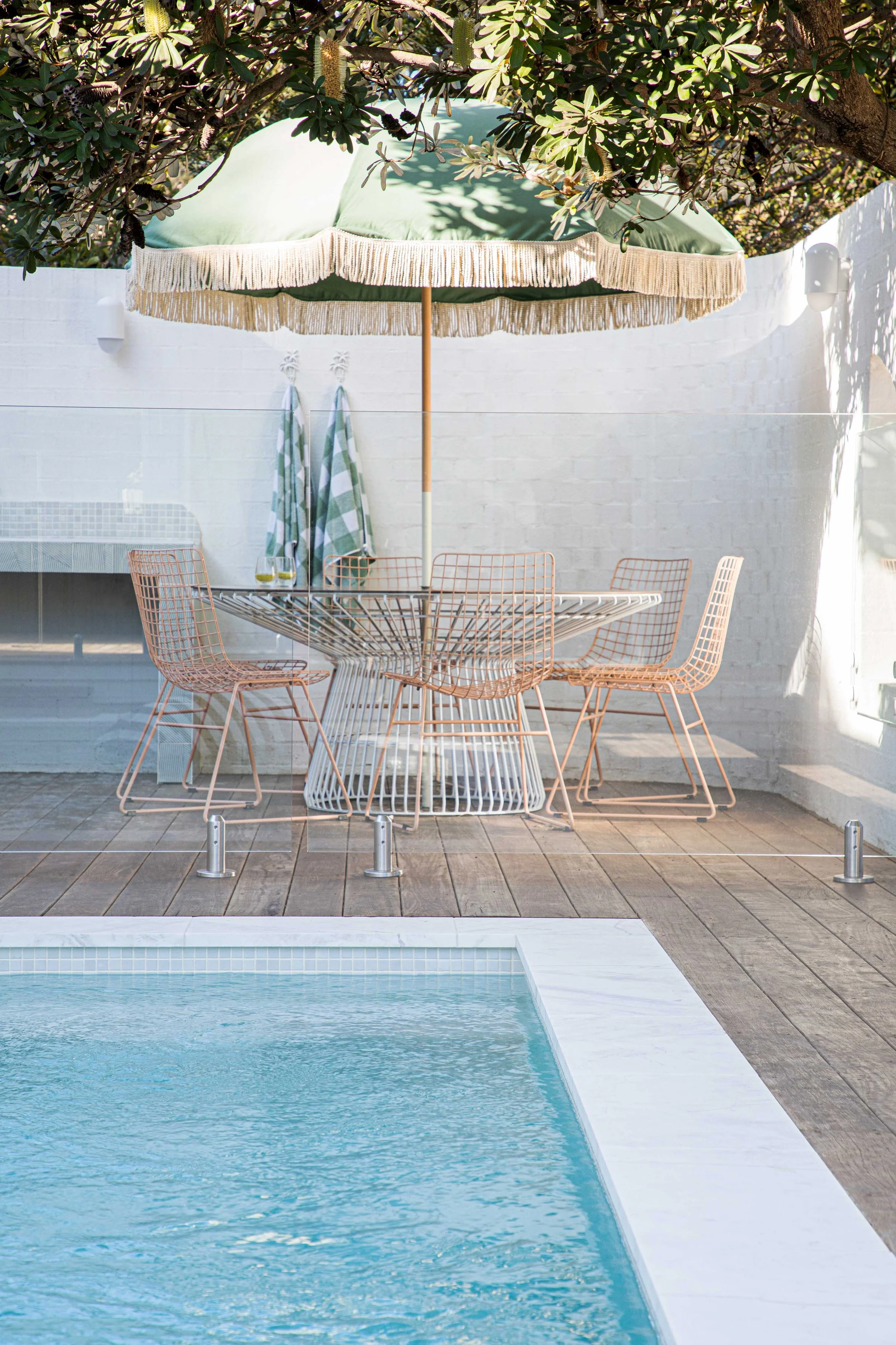 A view of a swimming pool with turquoise water, surrounded by a wooden deck. Behind the pool, there is a white brick wall with two outdoor lights and two striped towels hanging on hooks. Above the scene, a large green patio umbrella with a fringe ruf