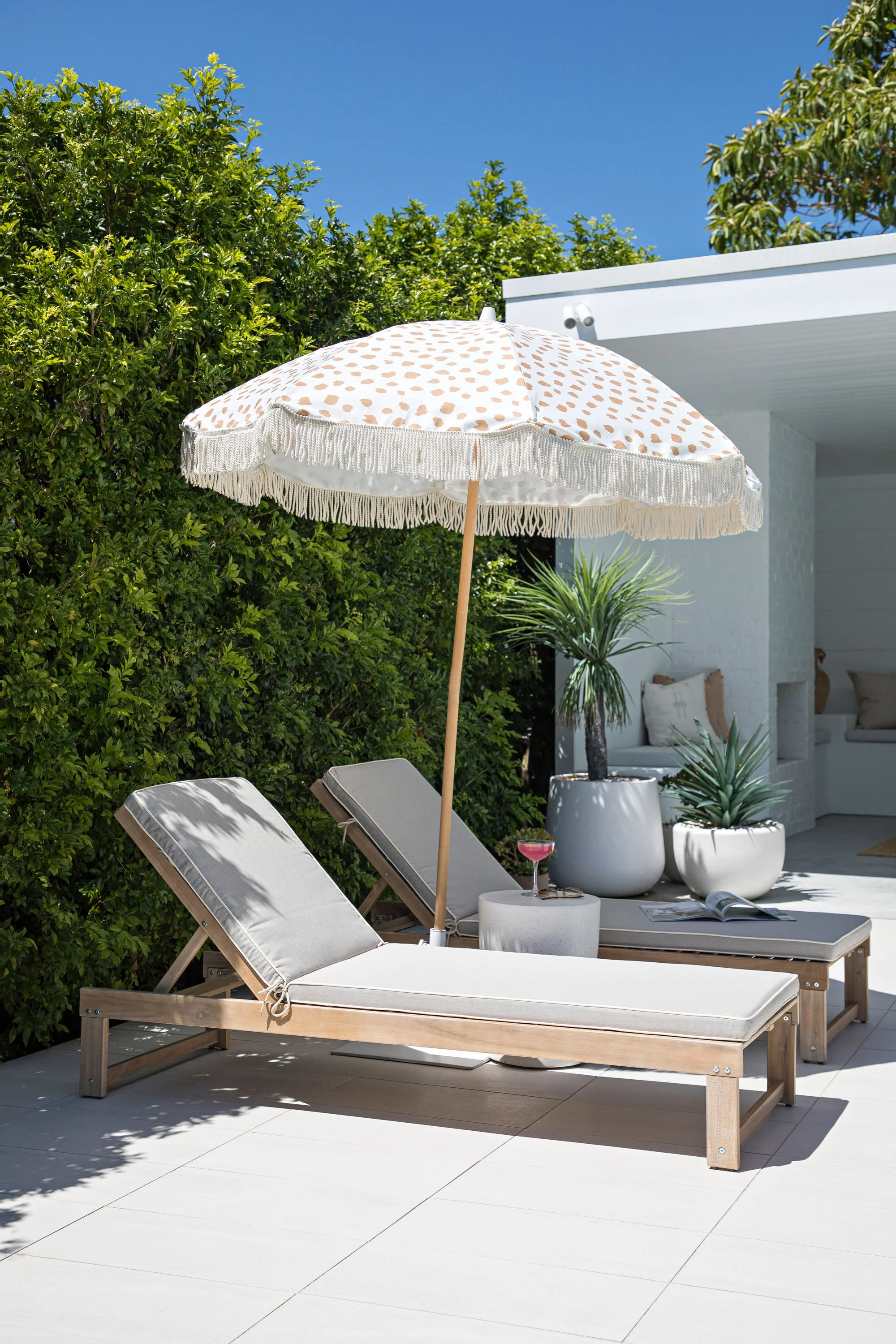 Outdoor patio area with two lounge chairs, a beach umbrella with a beige polka dot pattern and fringe, a glass of pink drink, a magazine, and potted plants under a clear blue sky.