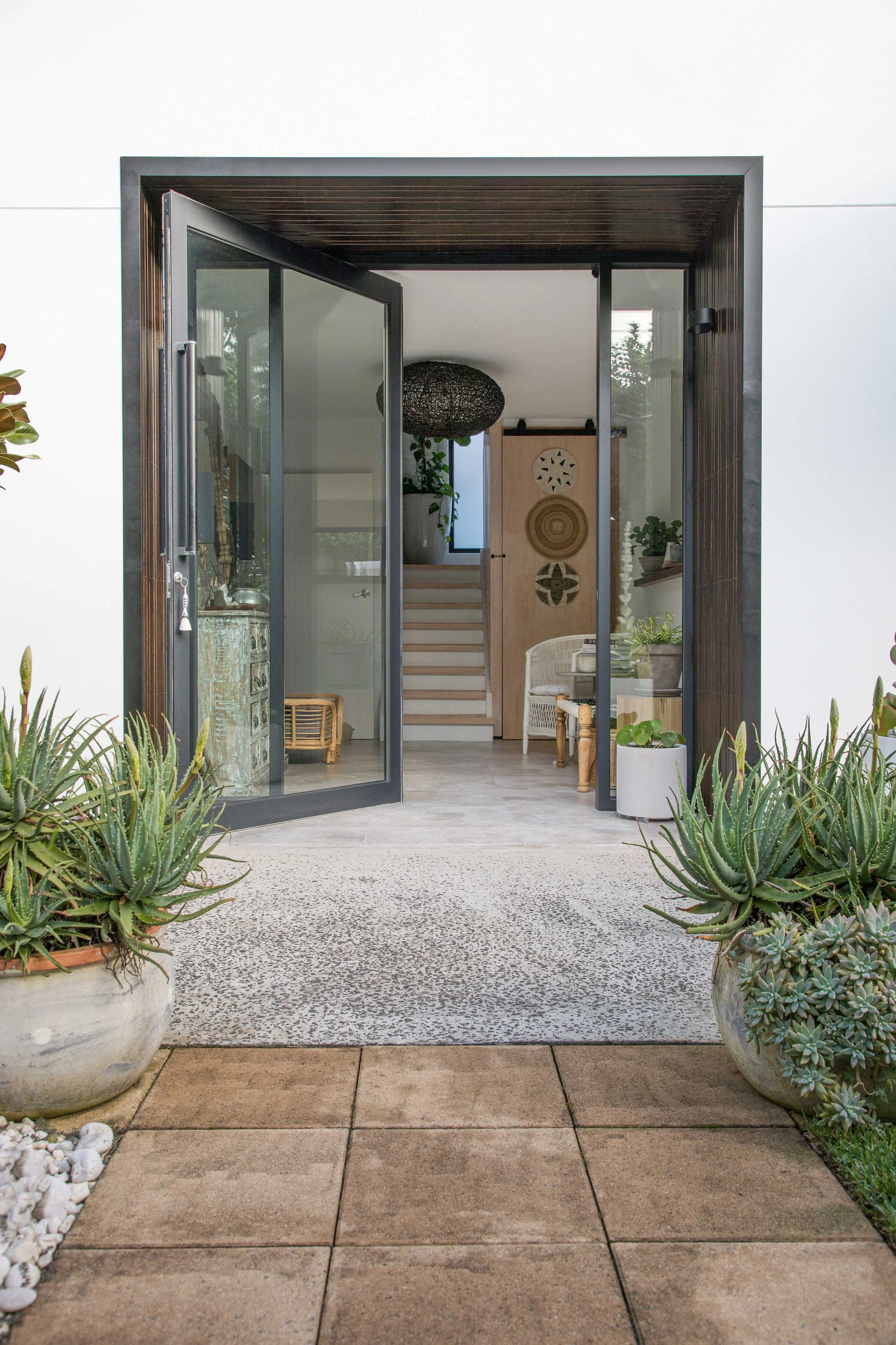Open glass door leading into a modern home, with an indoor seating area visible. Outside, there is a gravel pathway with large potted succulents and plants on both sides.