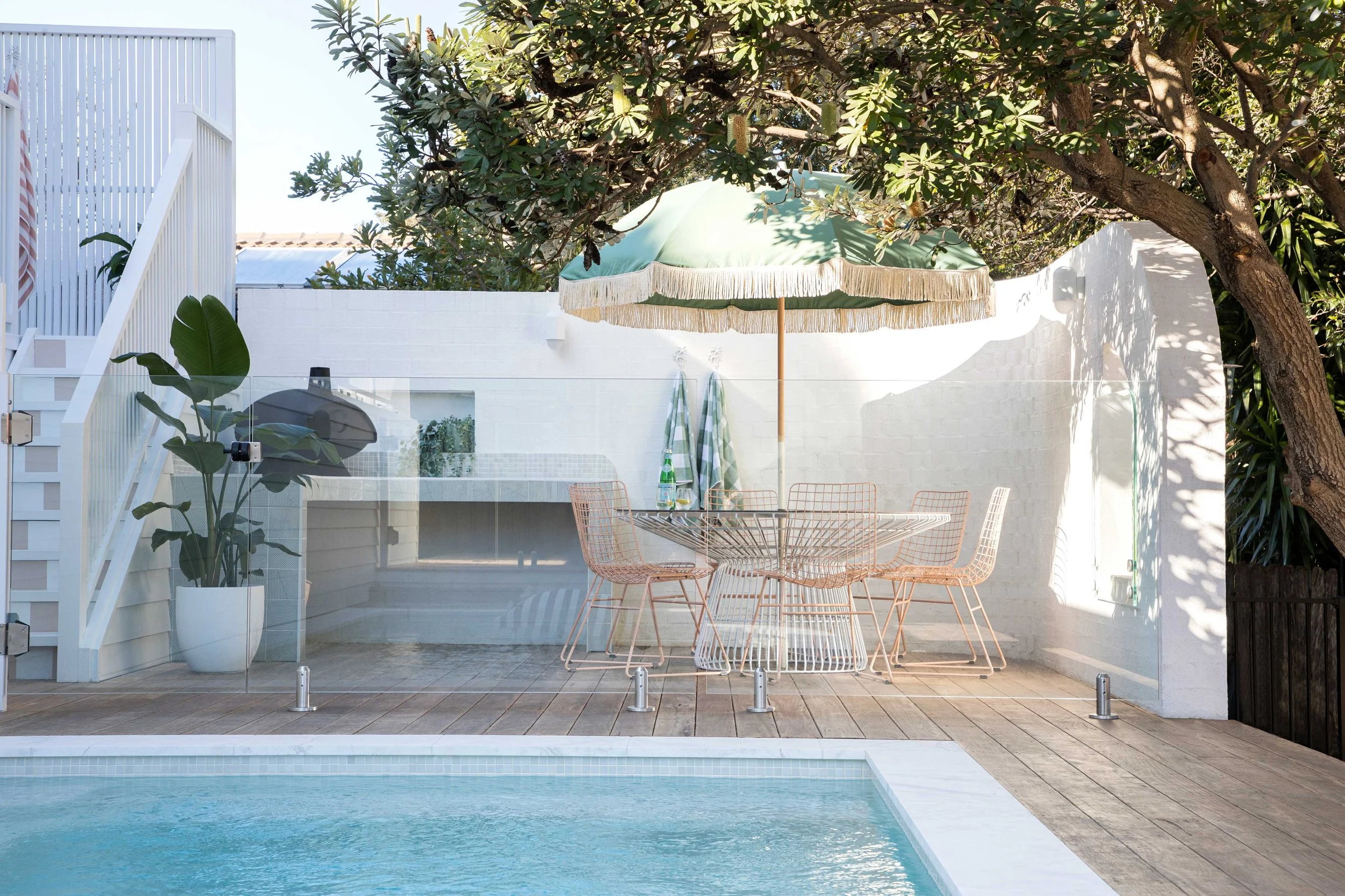 An outdoor patio area next to a swimming pool with a white wall, a glass table, and pink wire chairs. There is a large green umbrella providing shade and a tree with dense foliage overhead.