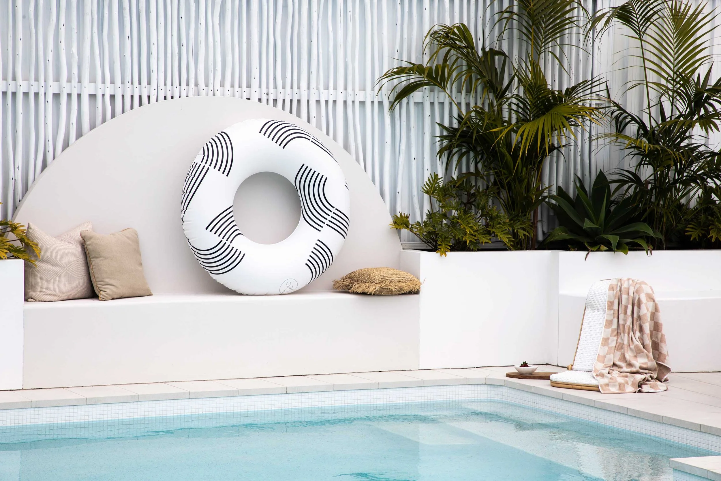 A private backyard swimming pool area with a white built-in bench, beige cushions, and a striped life preserver. Lush green plants and a white corrugated metal fence serve as the backdrop.