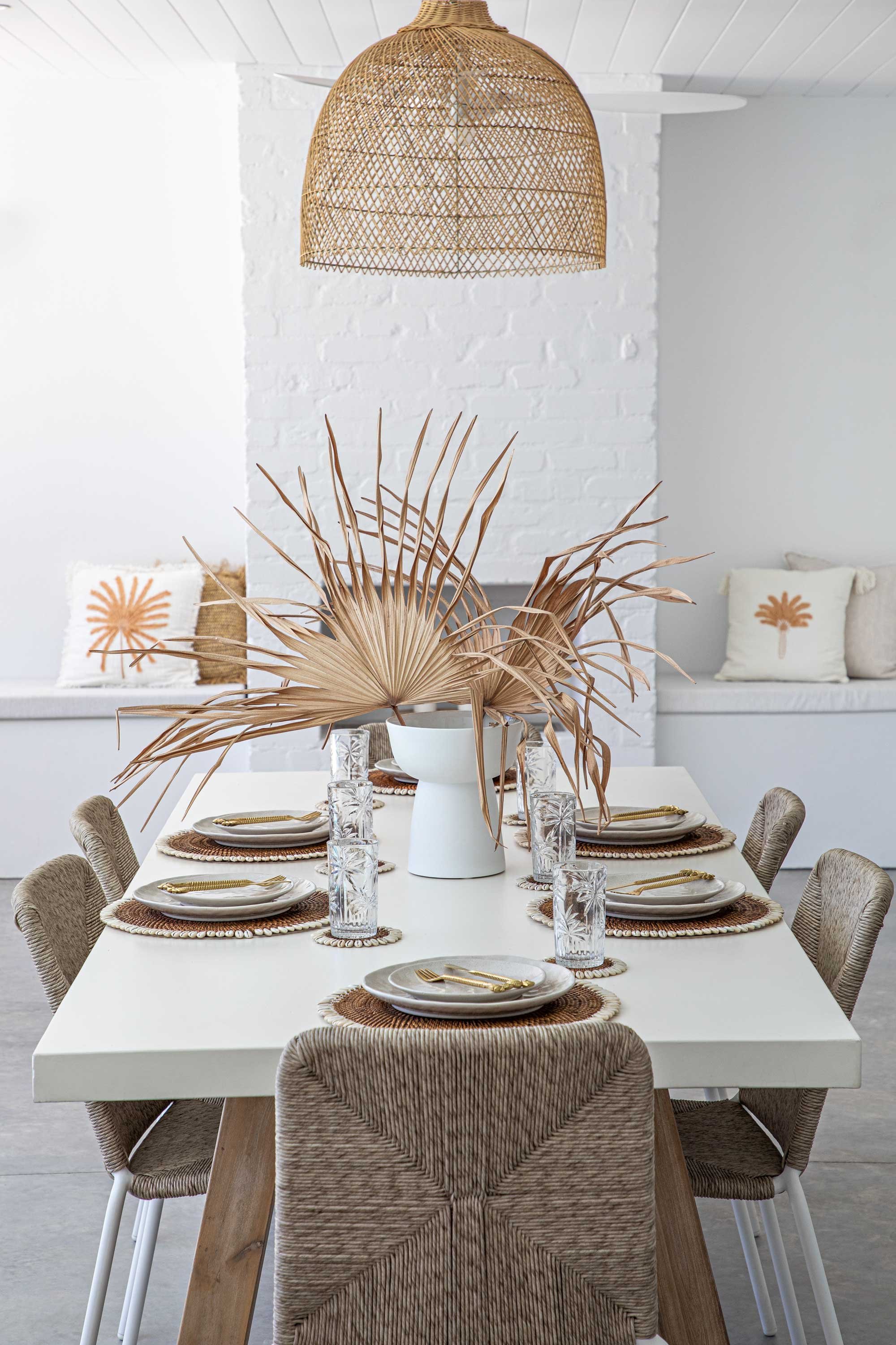 A dining table set for six with white plates, gold utensils, clear patterned glasses, and woven placemats. A white vase with dried palm leaves serves as a centerpiece. The table is surrounded by beige woven chairs. In the background, there are white 